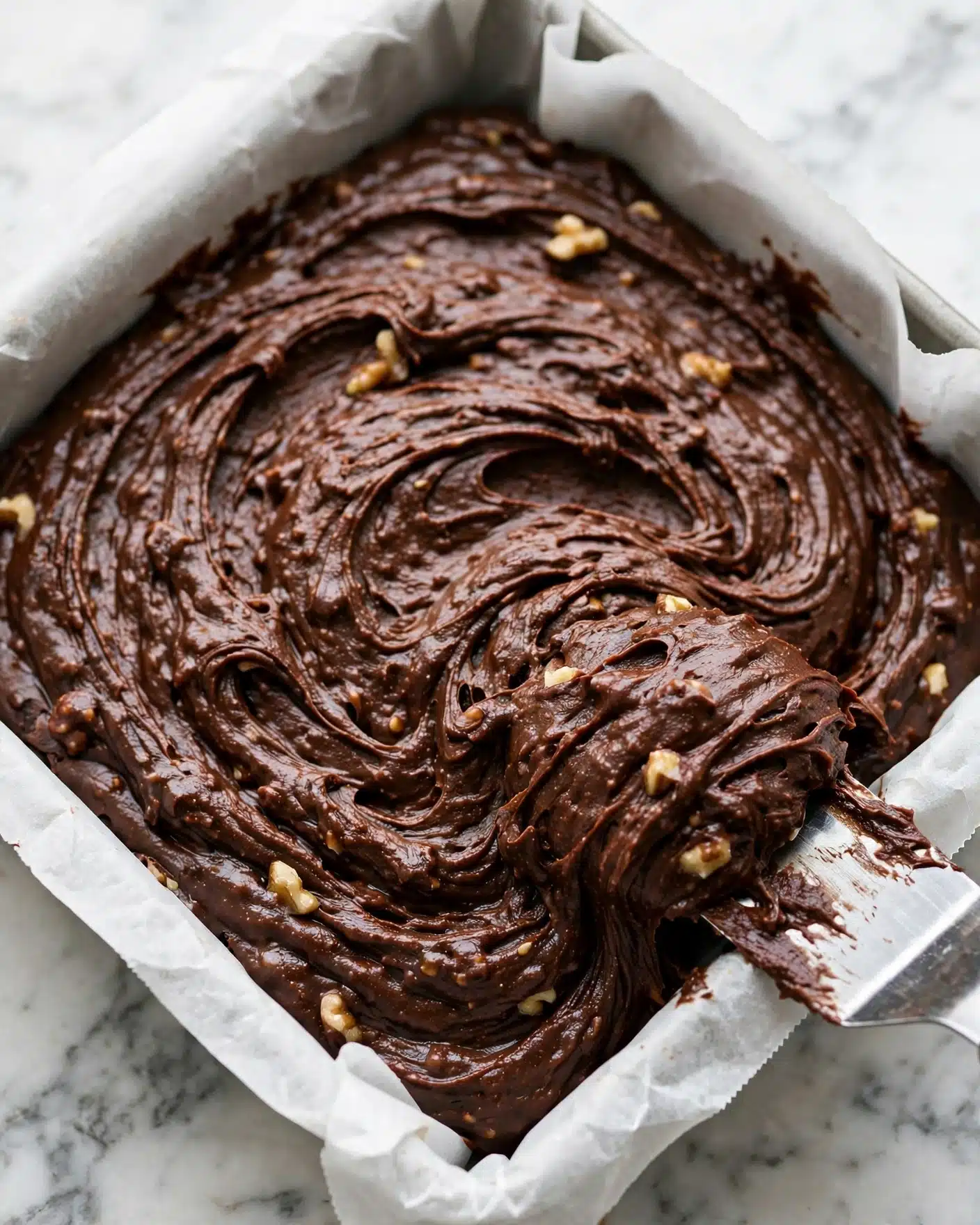 Easy Fudge Recipe 4 A square metal pan lined with white baking paper is filled with thick, rich dark chocolate batter that has small nut pieces scattered throughout. The batter's surface shows smooth, sweeping swirls and a textured, slightly glossy look, indicating it is well mixed but soft. A small metal spatula with some batter on its edge is being held by a woman's hand, touching the batter on the lower right corner. The pan rests on a white marbled surface. photo taken with an iphone --ar 4:5 --v 7