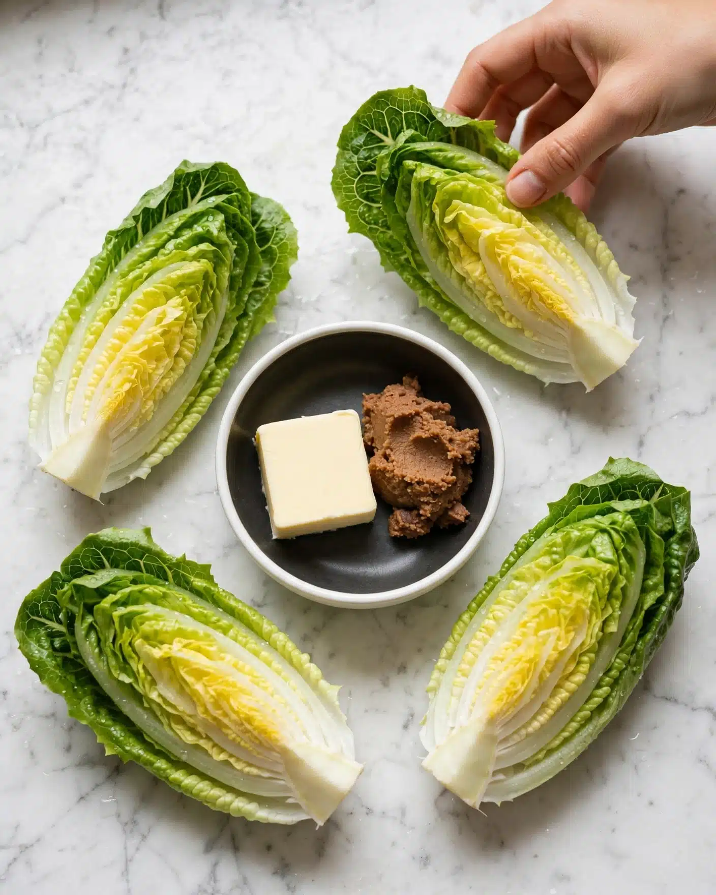 Miso Butter Roasted Cabbage Wedges Recipe 4 The image shows four wedges of green lettuce with light yellow centers placed on a white marbled surface, arranged around a white bowl with a dark inside. Inside the bowl, there is a square piece of light yellow butter and a small brown paste side by side. A woman's hand is holding one of the lettuce wedges from the top right corner. Photo taken with an iphone --ar 4:5 --v 7