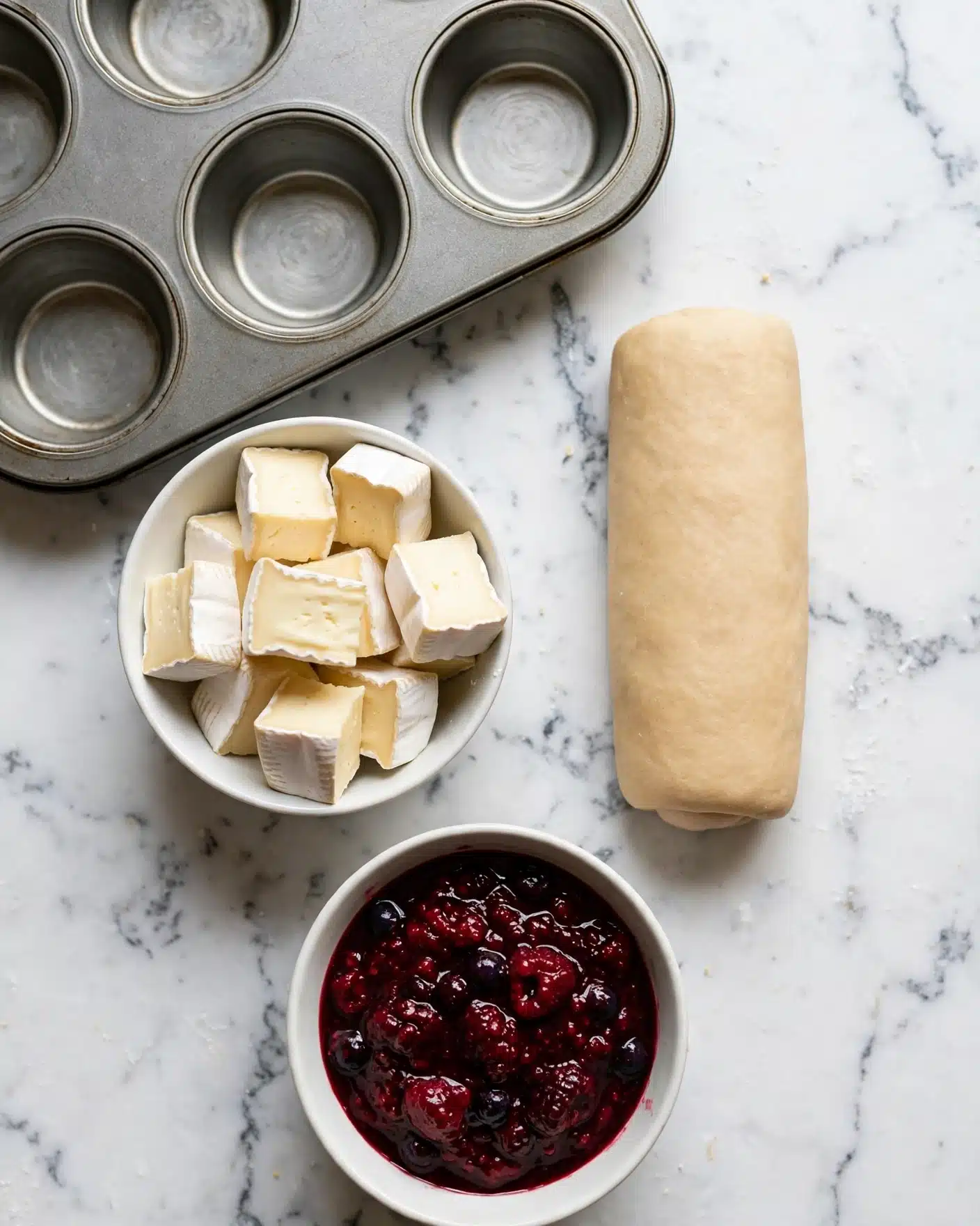Cranberry Brie Bites Recipe 4 The image shows a baking scene on a white marbled surface with a metal muffin tray at the top left. Below the tray, there is a small white bowl filled with light cream-colored cheese cubes, each with a soft texture and white rind edges. Near the bowl, to the right, there is a cylindrical roll of light tan dough with a smooth texture. At the bottom, a small white bowl contains a thick, dark red berry sauce with chunkier pieces visible, giving it a textured look. photo taken with an iphone --ar 4:5 --v 7