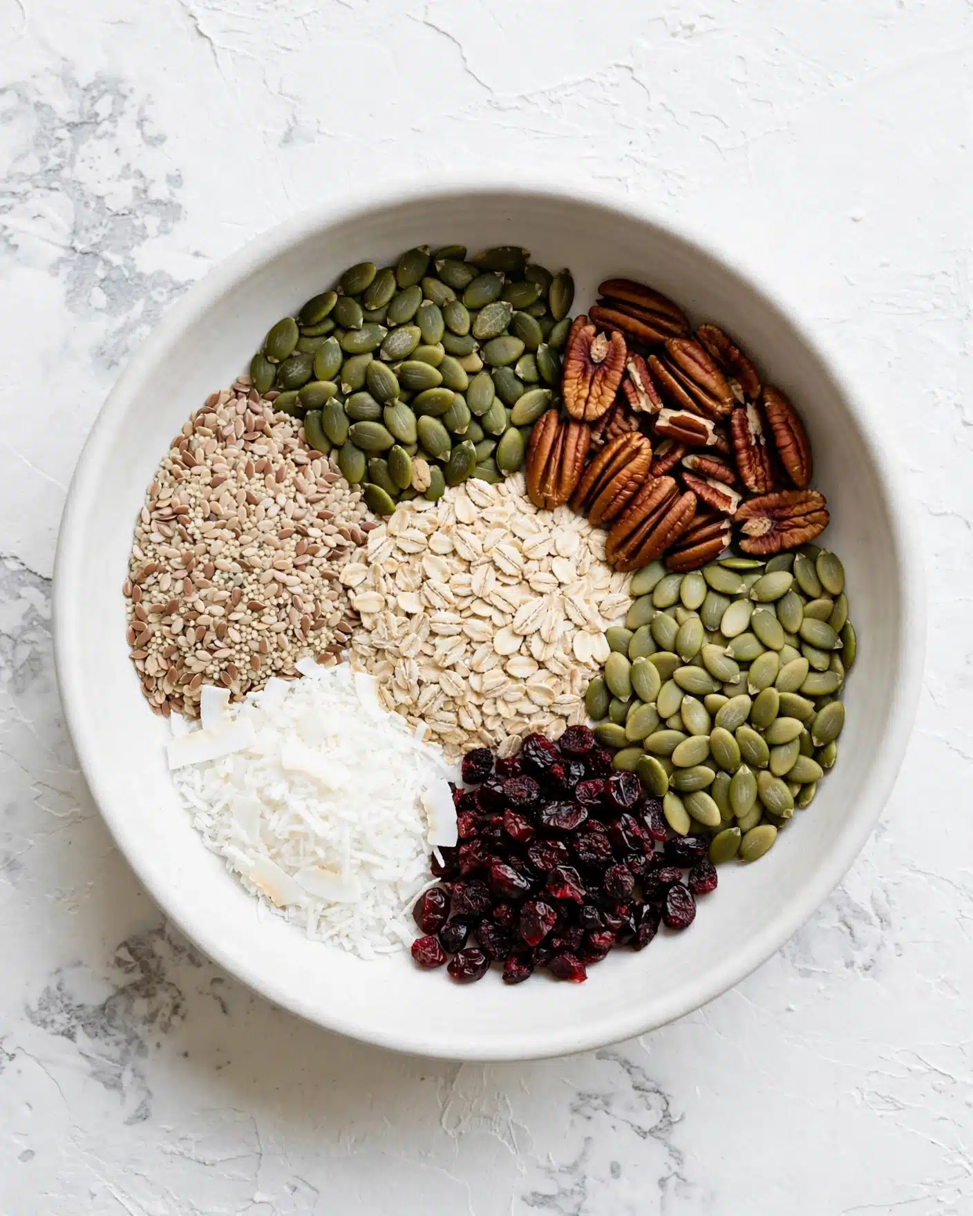 A white bowl sits on a white marbled textured surface, filled with six sections of different ingredients arranged like a colorful wheel. Starting from the top and moving clockwise, there are green pumpkin seeds, brown pecans, more pale green pumpkin seeds, small dark red dried berries, white coconut flakes, light beige flax seeds, and beige rolled oats in the center. Each section shows a rough texture and natural color, contrasting clearly with its neighbors. Photo taken with an iphone --ar 4:5 --v 7