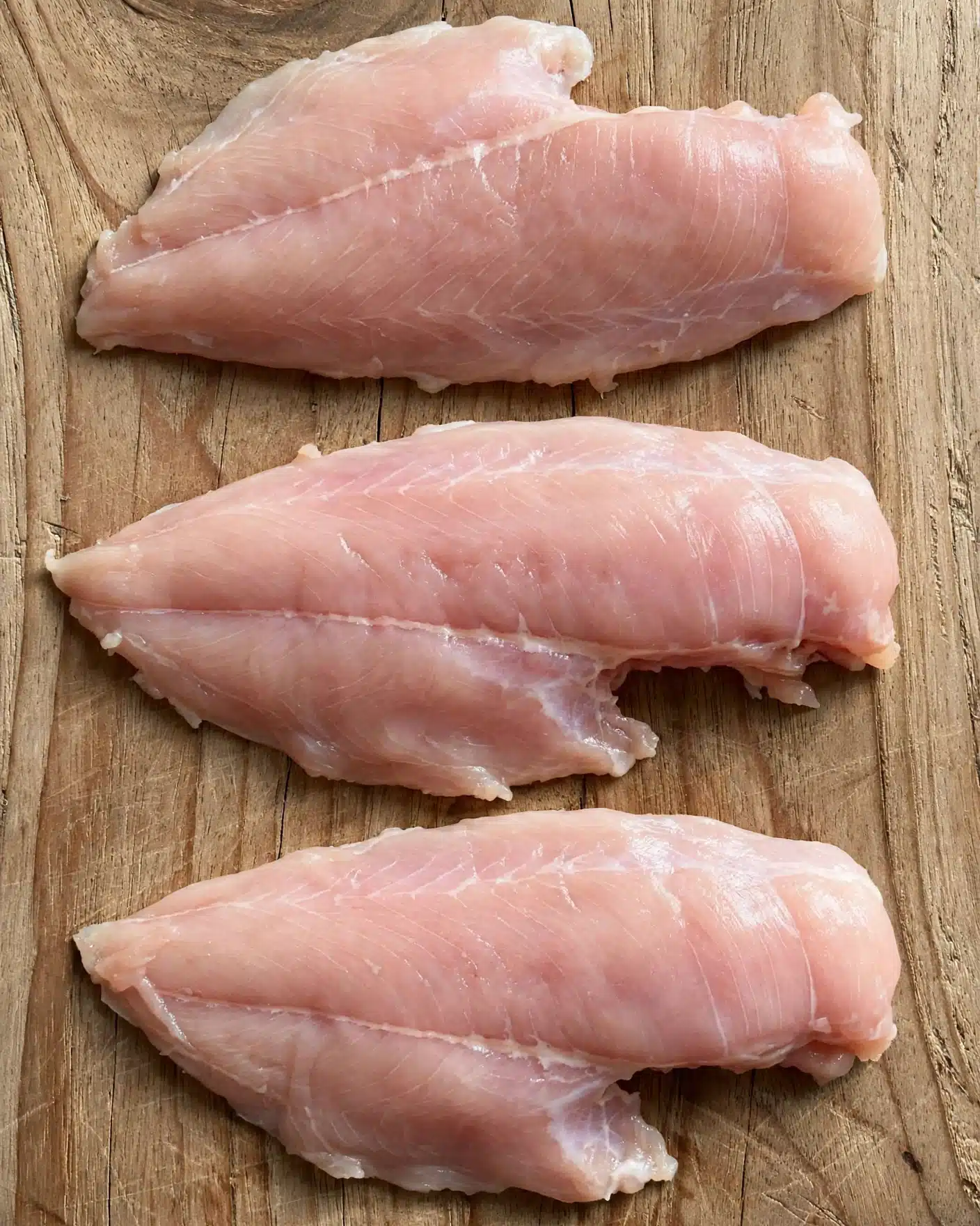 Three raw chicken fillets lie flat on a wooden surface with visible grain texture. Each fillet has a smooth, slightly shiny light pink surface with subtle variations in color and faint muscle lines. The edges are soft and slightly irregular. The photo taken with an iphone --ar 4:5 --v 7