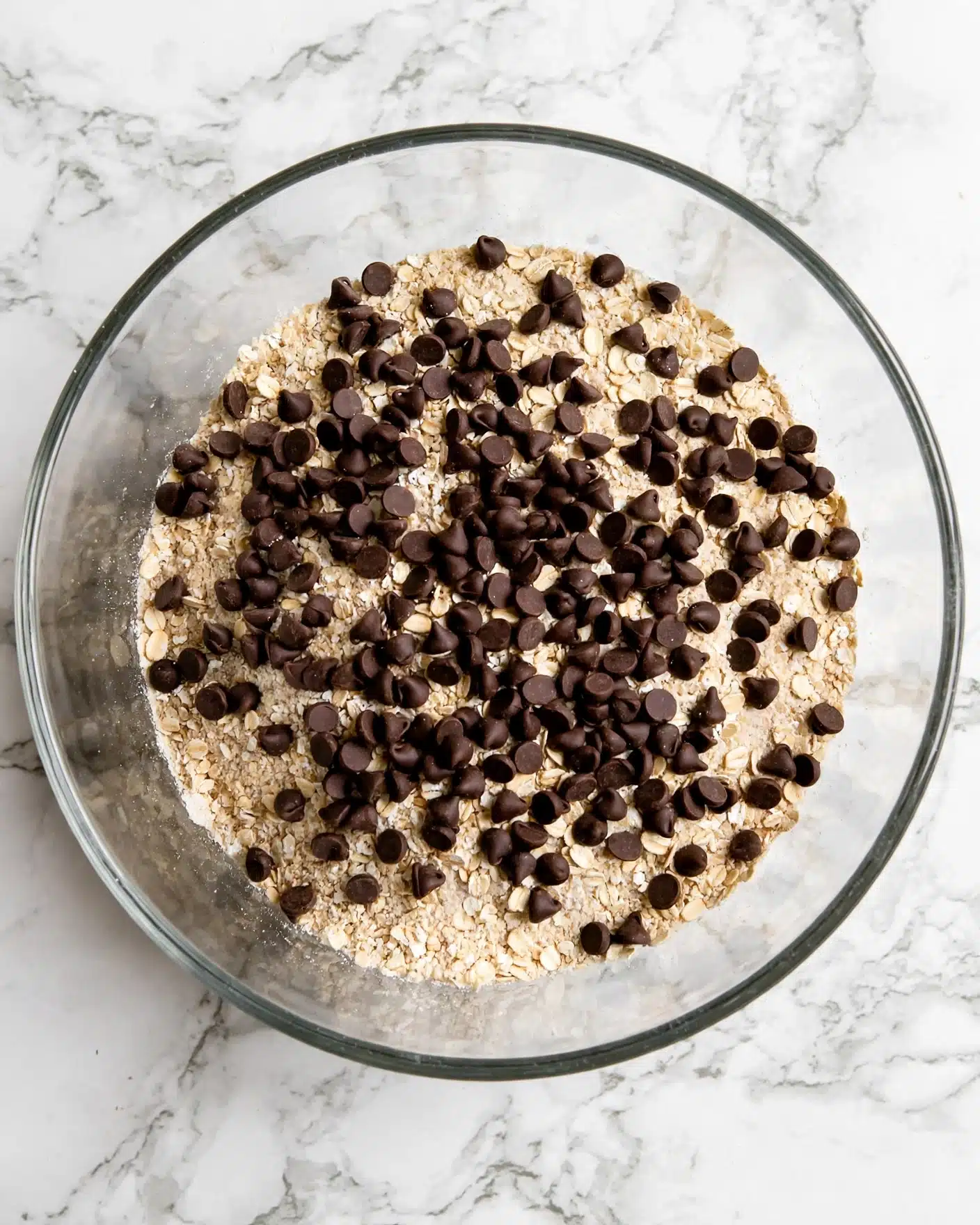 A clear glass bowl sits on a white marbled surface, filled with a mixture of light-colored dry ingredients forming the base layer with a slightly rough texture. On top, there is a large, uneven layer of small, dark brown chocolate chips scattered randomly, creating a contrast against the lighter mixture below. The bowl is shown from above with all layers visible clearly. Photo taken with an iphone --ar 4:5 --v 7