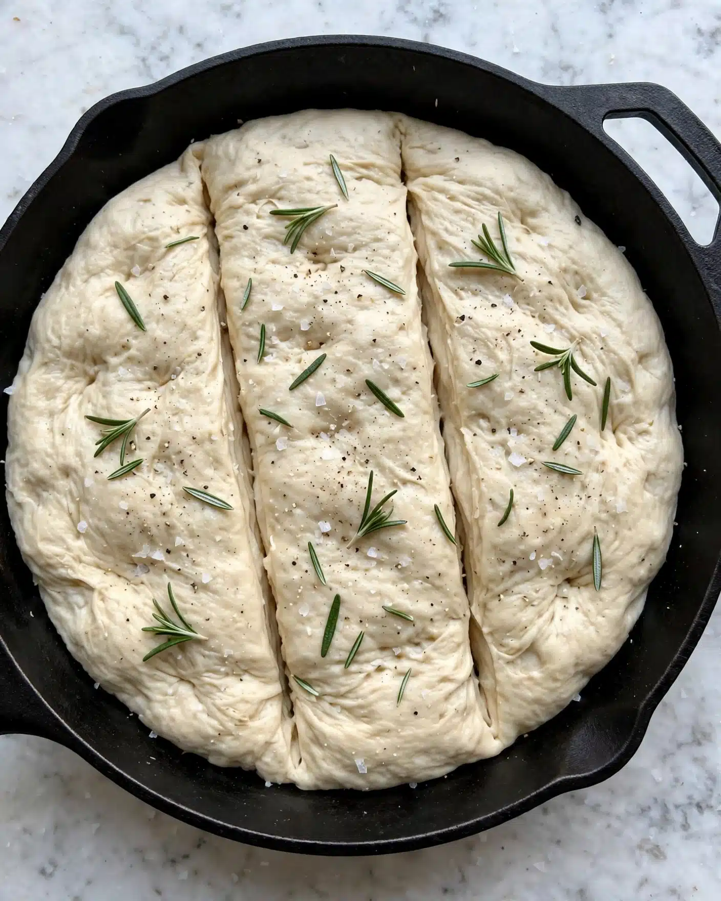 A round, thick layer of pale dough with a soft, slightly wrinkled texture fills a black cast iron pan. The top surface of the dough is sprinkled with small green rosemary leaves and a light dusting of coarse salt and pepper. The dough has two long shallow cuts running from top to bottom, dividing it into three parts. The pan rests on a white marbled surface. photo taken with an iphone --ar 4:5 --v 7