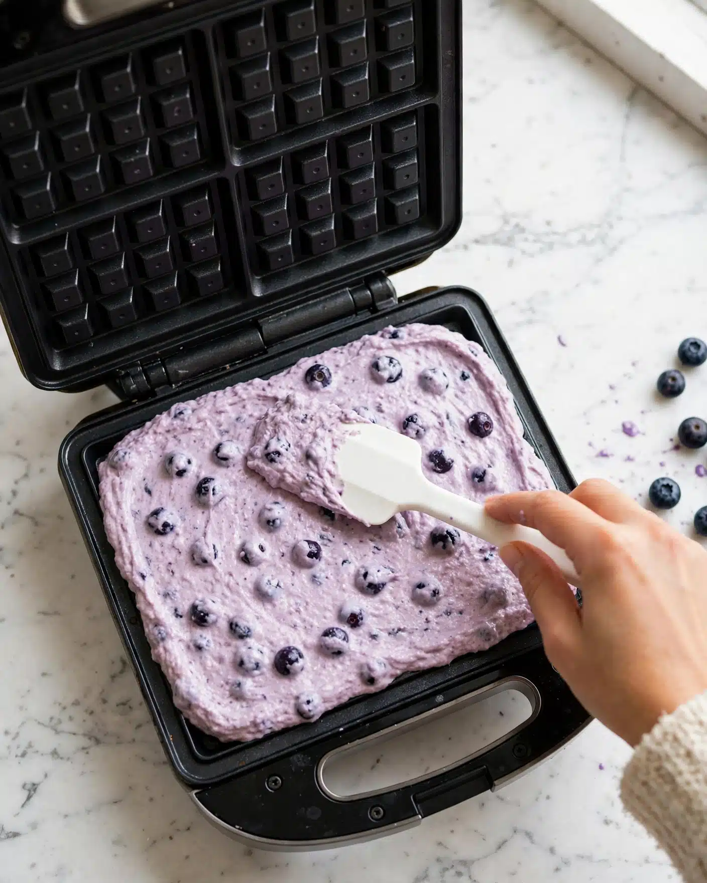 The image shows a black waffle maker open on a white marbled surface with a white spatula spreading a thick, light purple batter containing visible dark purple blueberries evenly across the square mold. The batter covers the entire bottom plate of the waffle maker, showing a creamy texture mixed with the fruit. A woman's hand holds the spatula, gently smoothing the mixture. Photo taken with an iphone --ar 4:5 --v 7