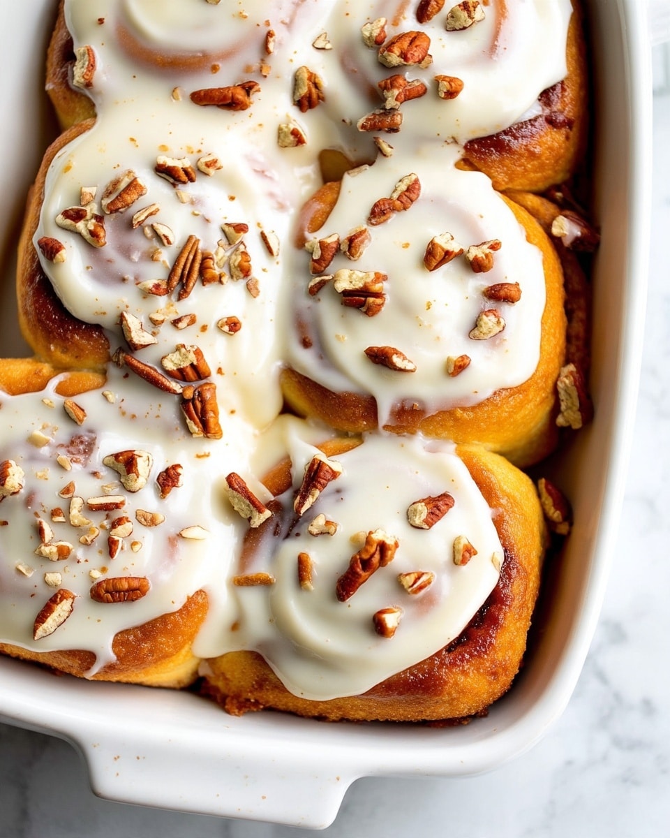 The image shows a white rectangular baking dish filled with cinnamon rolls arranged in close layers. Each cinnamon roll has visible swirls of dark brown cinnamon filling that contrast with the warm golden-brown dough. The rolls are topped generously with smooth, white icing that drips along their sides, and small pieces of pecans are scattered on top and between the rolls, adding a crunchy texture. The dish is set on a white marbled surface with a hint of wooden tones nearby. Photo taken with an iphone --ar 4:5 --v 7