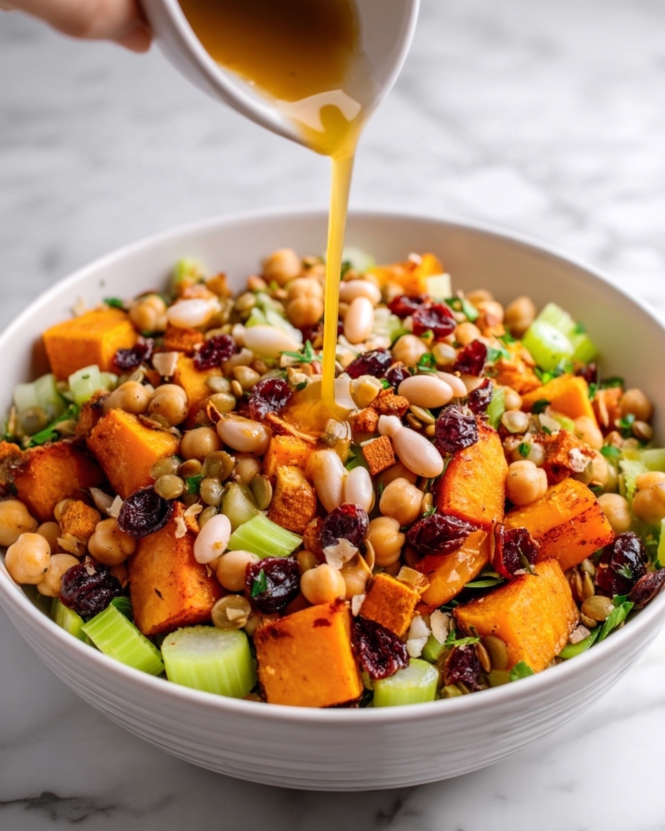 A white bowl filled with a colorful salad featuring large orange roasted sweet potato cubes as the base layer, topped with light green celery pieces and beige chickpeas scattered throughout. Small white beans and lentils add texture, while bits of fresh green herbs sprinkle across the surface. Dark red dried cranberries are spread on top, adding contrast to the mix. A woman's hand is pouring a golden liquid dressing from above, creating a streak going down into the salad. The bowl sits on a white marbled surface. photo taken with an iphone --ar 4:5 --v 7