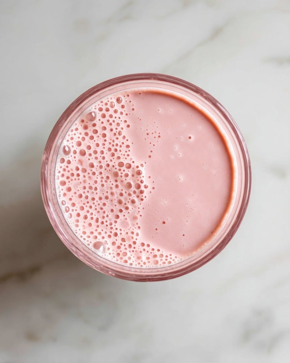 A top view of a clear glass filled with a light pink smoothie. The smoothie has a smooth texture with small bubbles spread across the surface. The glass has a slight ripple design along the rim. It is placed on a white marbled surface. photo taken with an iphone --ar 4:5 --v 7