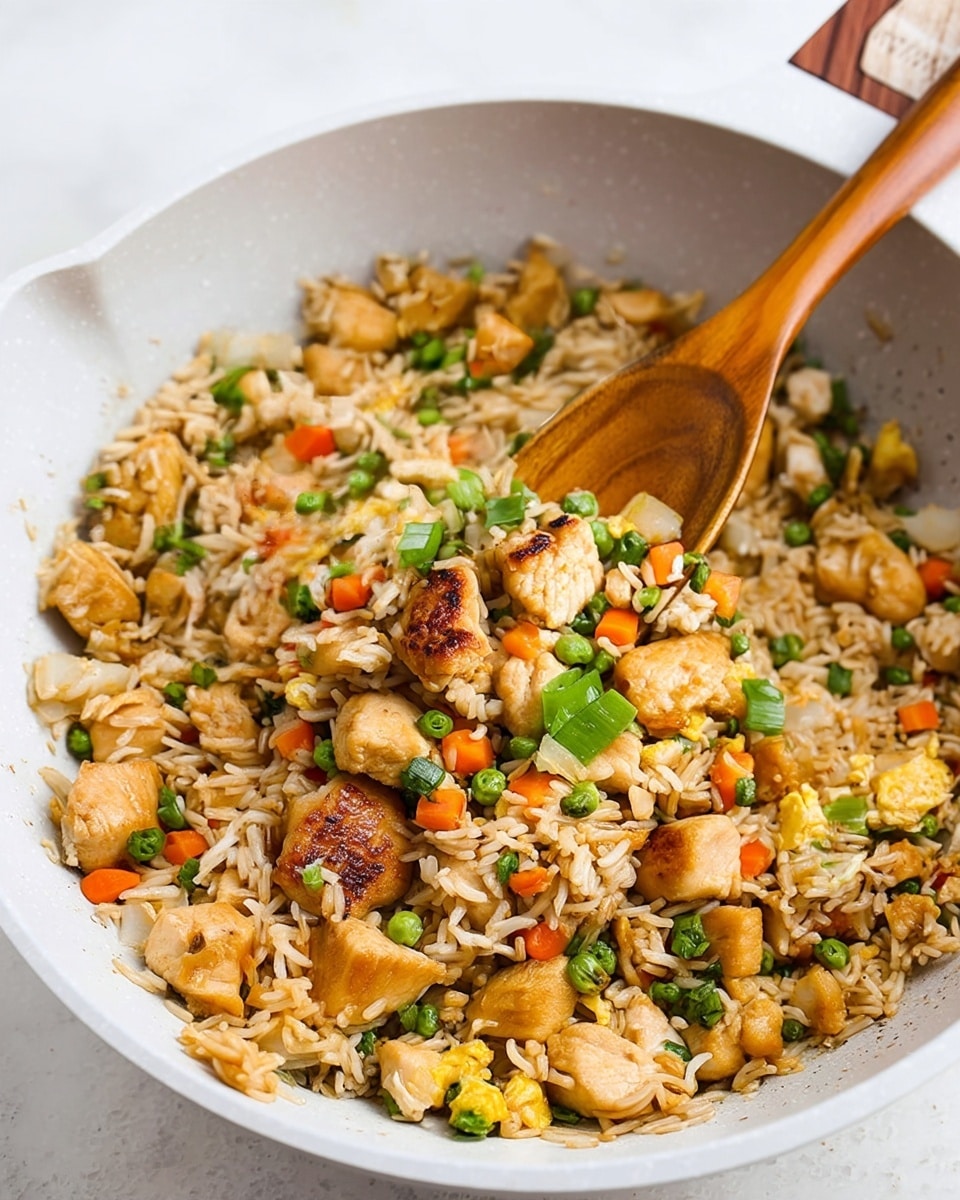 A white bowl with a brown rim filled with fried rice showing several layers: light brown fried rice grains mixed with small bright orange carrot cubes, green peas, light yellow scrambled egg pieces, white onion chunks, and medium brown cooked chicken pieces. On top, there are bright green sliced scallions. Two dark brown chopsticks rest inside the bowl on the left side. The bowl sits on a white marbled surface with a small bowl of green herbs blurred in the background. Photo taken with an iphone --ar 4:5 --v 7