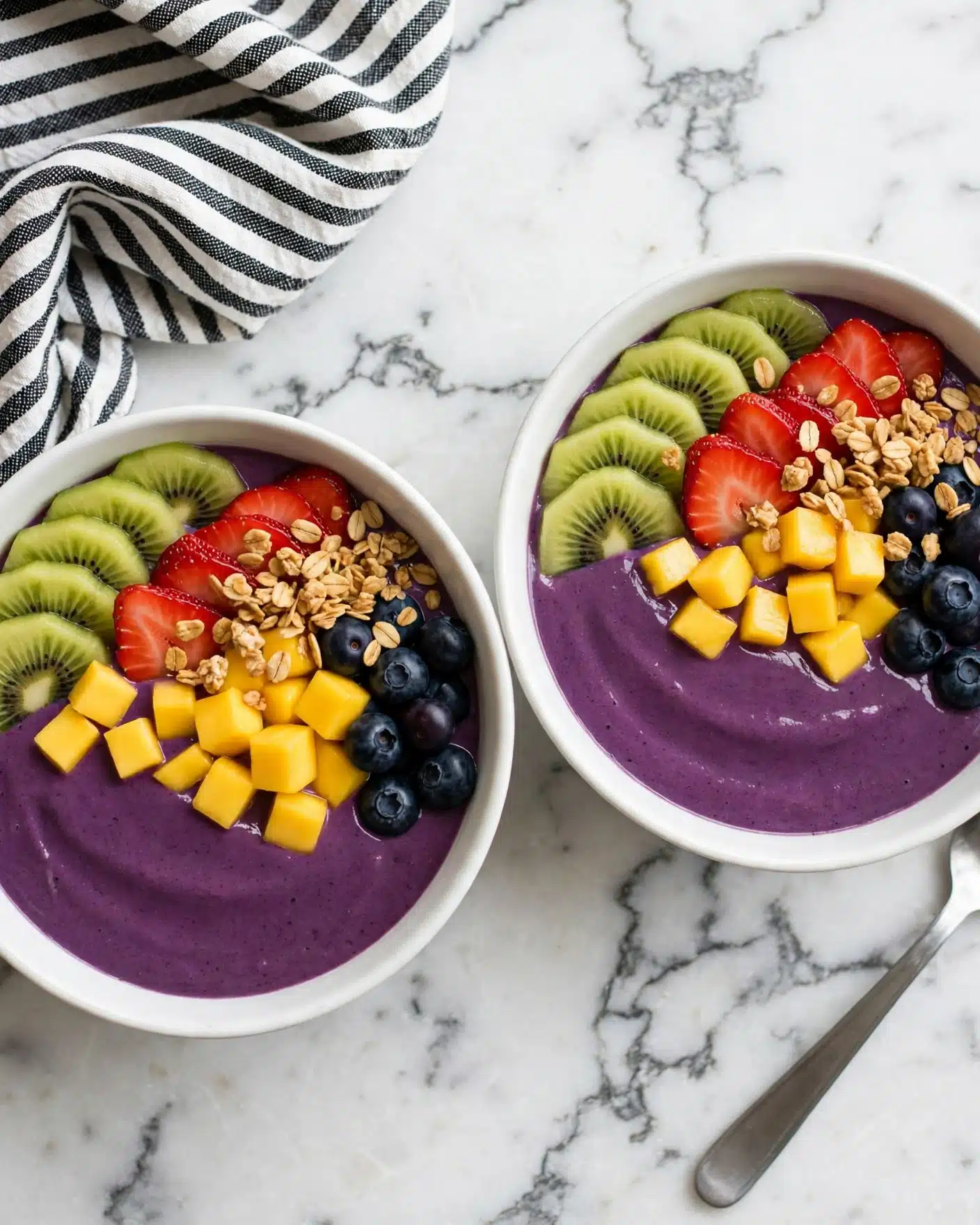 A white bowl filled with a thick purple smoothie forms the base layer. On top, there are slices of bright green kiwi arranged in a curved row on one side. Next to the kiwi, there are bright red strawberry slices lined up neatly. Beside the strawberries, small yellow mango cubes are placed. Blueberries with a deep blue color sit next to the mango pieces. Scattered over and around the blueberries and mango is a layer of golden-brown granola with visible oats and nuts. The bowl is on a white marbled surface with a black and white striped cloth slightly visible in the background. A silver spoon rests inside the bowl on the right side. Photo taken with an iphone --ar 4:5 --v 7