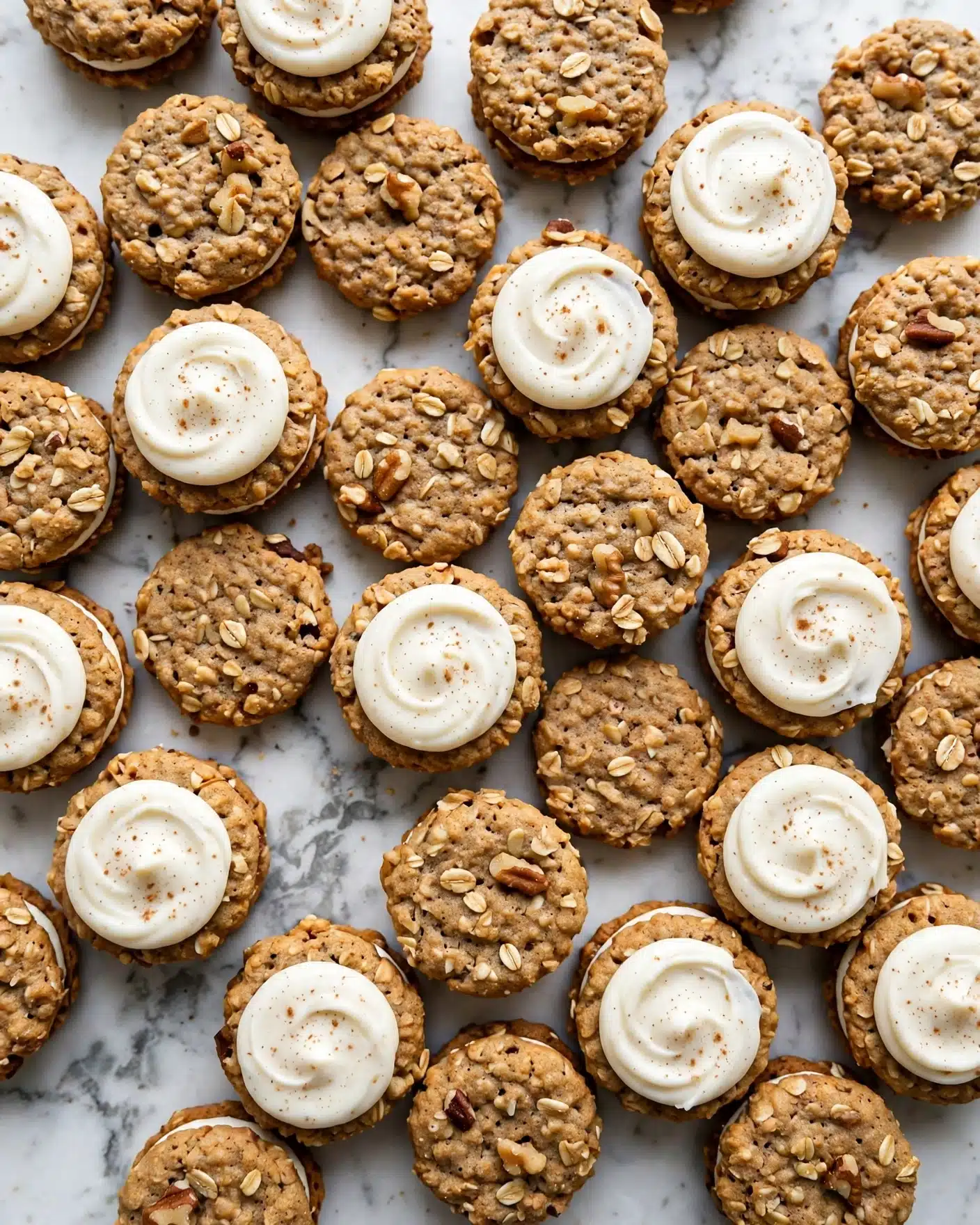 The image shows many small round cookies laid out on a white marbled surface. Each cookie is golden brown with a slightly rough texture and visible oats or nuts. About half of the cookies have a dollop of smooth, creamy white frosting with tiny specks of spice on top, swirled in a circular pattern. The cookies without frosting show a more textured, somewhat uneven surface. The arrangement is close together, filling the whole frame evenly. Photo taken with an iphone --ar 4:5 --v 7