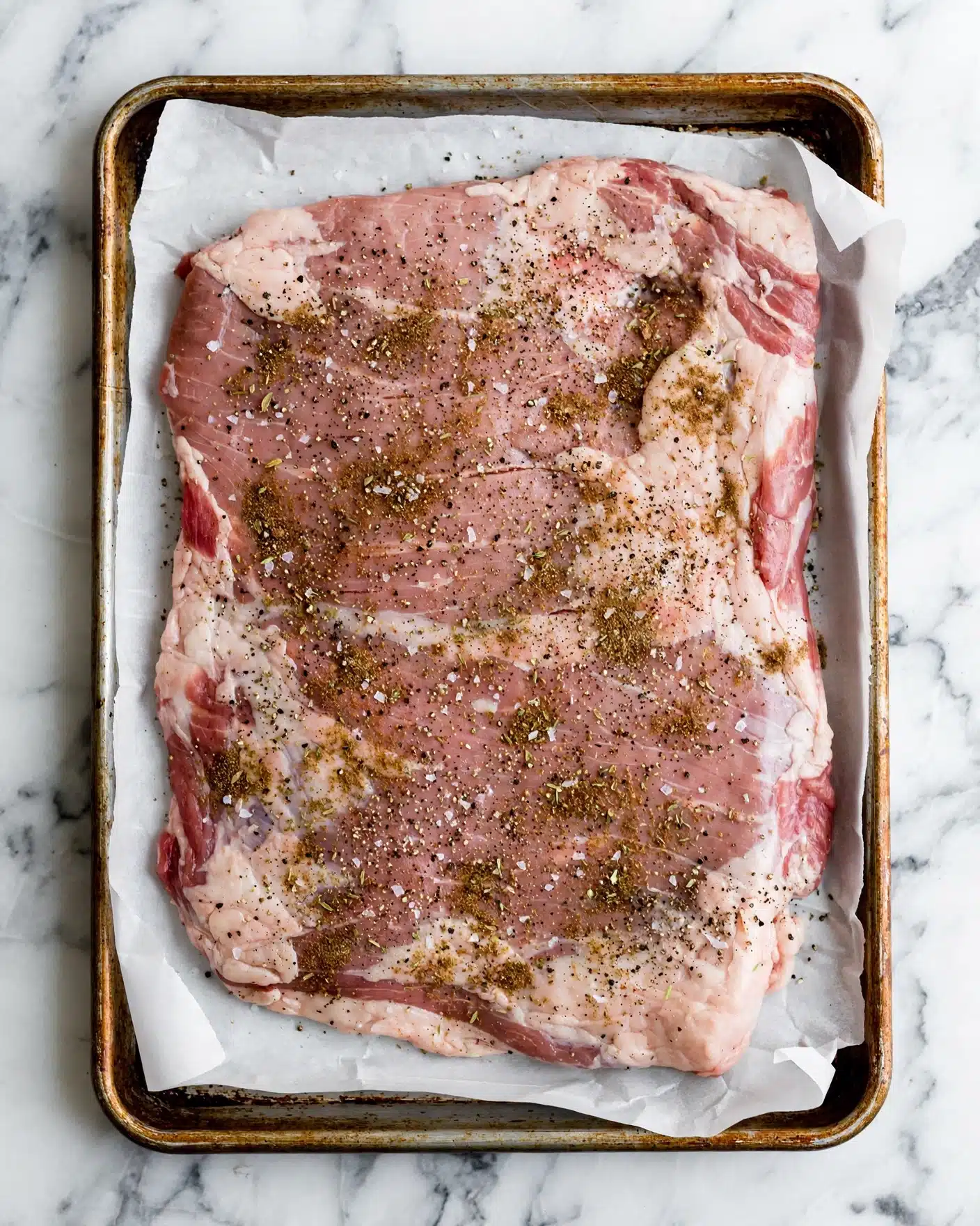 Juicy Oven Roasted Turkey Breast with Brown Sugar and Butter Recipe 4 A raw piece of meat lies on a white parchment paper on a metal baking tray with a worn, rusty edge. The meat is large and flat, mostly pale pink with areas of light pink and white fat along the edges and throughout. It is covered in a seasoning mix of coarse salt, black pepper, and other brownish spices, sprinkled unevenly over the surface. The baking tray sits on a white marbled texture background photo taken with an iphone --ar 4:5 --v 7