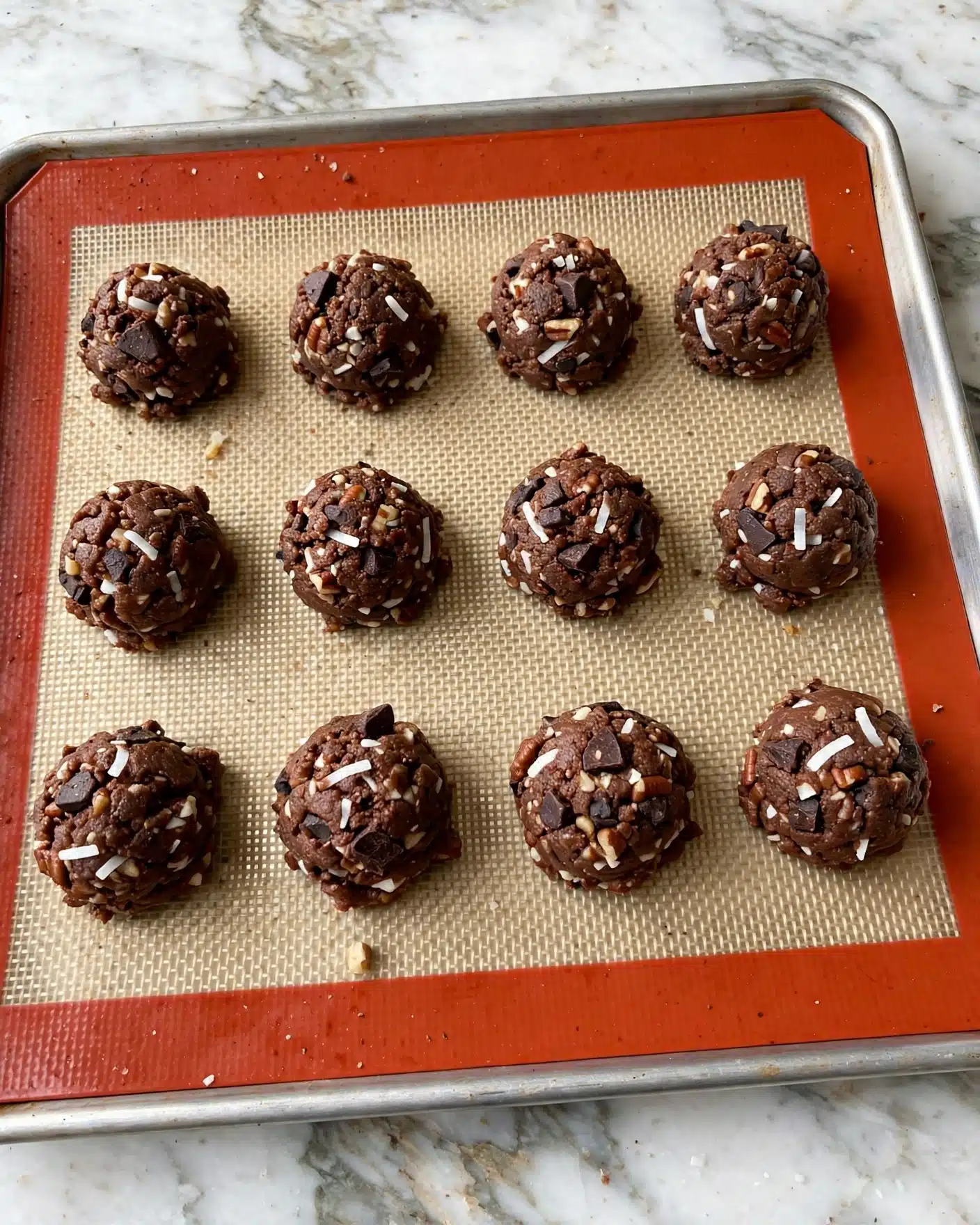 German Chocolate Cookies Recipe 5 Twelve small, round balls of dark brown chocolate cookie dough sit evenly spaced on a baking sheet lined with a red-bordered silicone mat, which is on a white marbled surface. Each dough ball is textured with small chunks of white coconut flakes and lighter brown pieces that look like nuts, along with darker chocolate bits scattered throughout. The dough balls have a slightly rough, chunky look and are arranged in three rows of four across the mat. Photo taken with an iphone --ar 4:5 --v 7