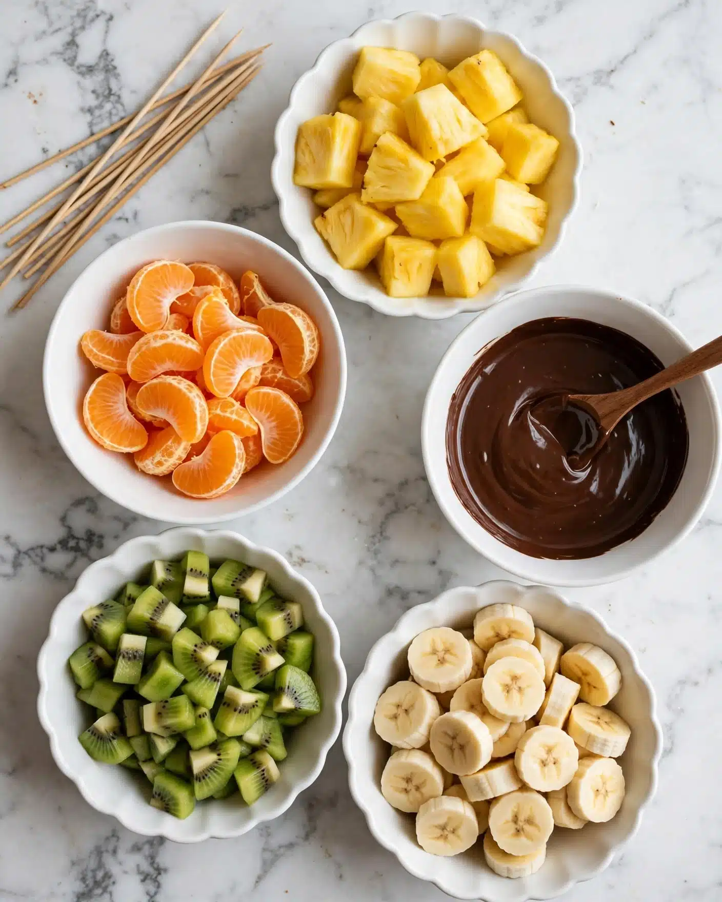 Rainbow Fruit Skewers with Dark Chocolate Drizzle Recipe 4 The image shows five white bowls arranged in a circle on a white marbled surface, each with different ingredients inside. Starting from the top right, there is a scalloped bowl filled with yellow pineapple chunks. To the right, a plain bowl holds dark melted chocolate with a spoon resting inside. Below it, a scalloped bowl contains cut banana pieces. To the bottom left, another scalloped bowl has green kiwi chunks with black seeds. At the top left, a plain bowl holds bright orange mandarin segments. Several wooden skewers lay on the top left side of the surface. photo taken with an iphone --ar 4:5 --v 7