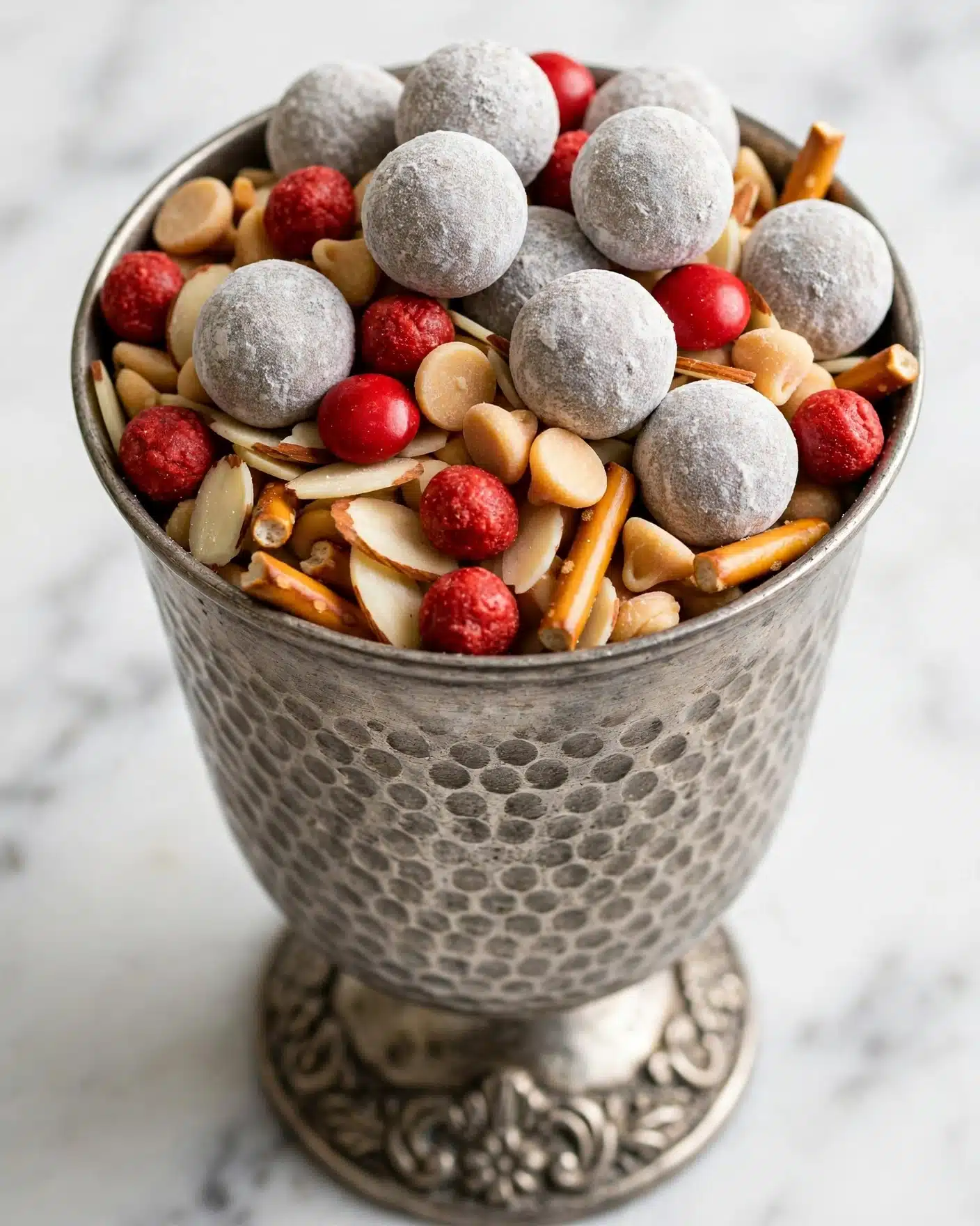 Werewolf Kibble Recipe 4 A close-up view of a metal goblet filled with a snack mix showing multiple layers, including round, grayish balls dusted with powder on top, scattered bright red round pieces, small beige butterscotch-colored chips, thin almond slices, and small pretzel sticks. The goblet has a textured surface with a decorative base, and the background is a white marbled texture. photo taken with an iphone --ar 4:5 --v 7