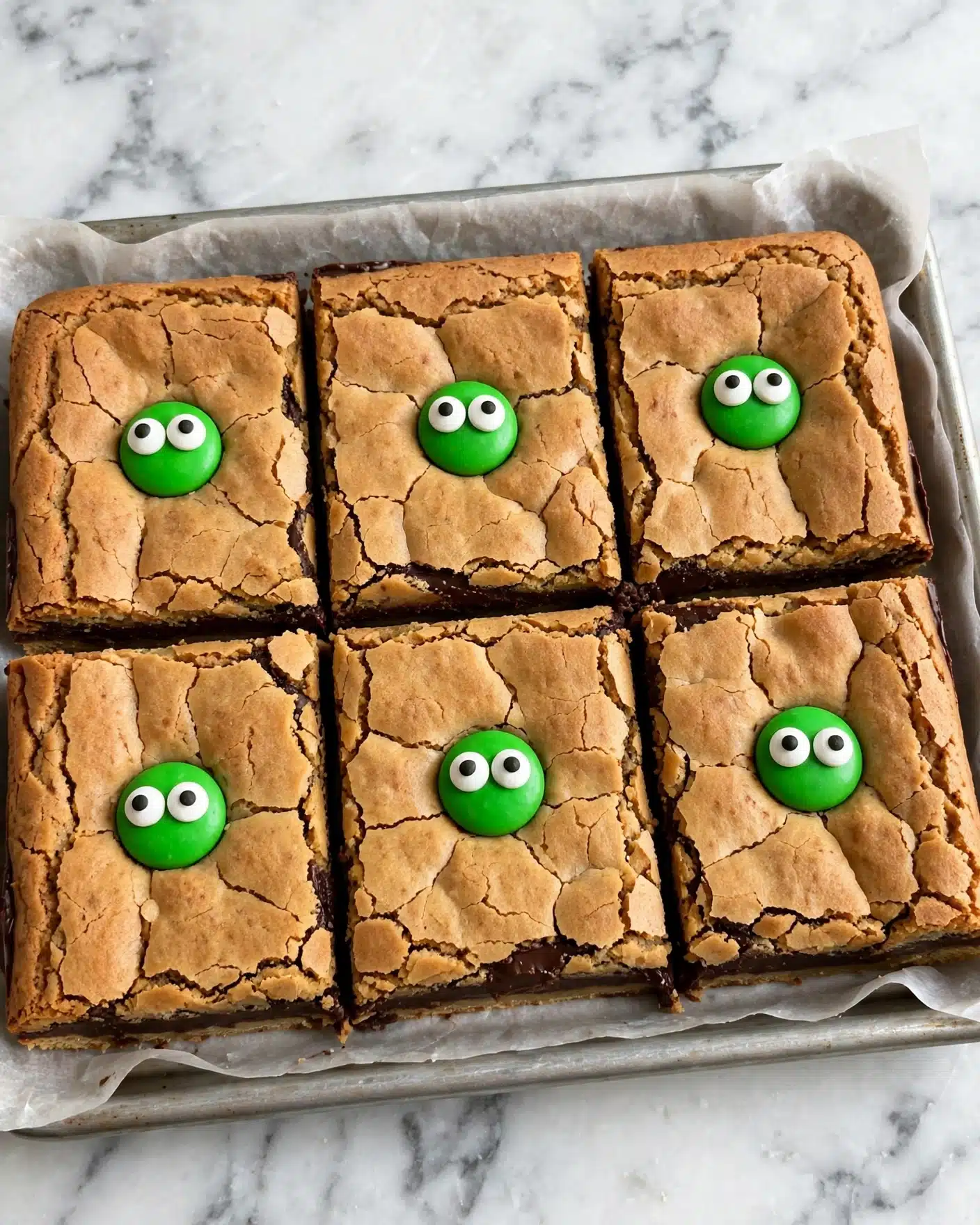 Monster Peanut Butter Cookie Bars Recipe 5 The image shows a grid of twelve square cookie bars placed on white parchment paper over a white marbled surface. Each bar has a golden-brown, slightly cracked top layer with a soft texture. Embedded in the bars are circular green frosting blobs decorated with one or two round candy eyes, which have white and black parts, giving a playful monster look. Additional candy eyes are scattered throughout the cookie bars, some partially hidden under the cookie crust. On the left side of the white marbled surface are many loose candy eyes, and a white cloth with black polka dots is partially visible in the top right corner of the image. Photo taken with an iphone --ar 4:5 --v 7