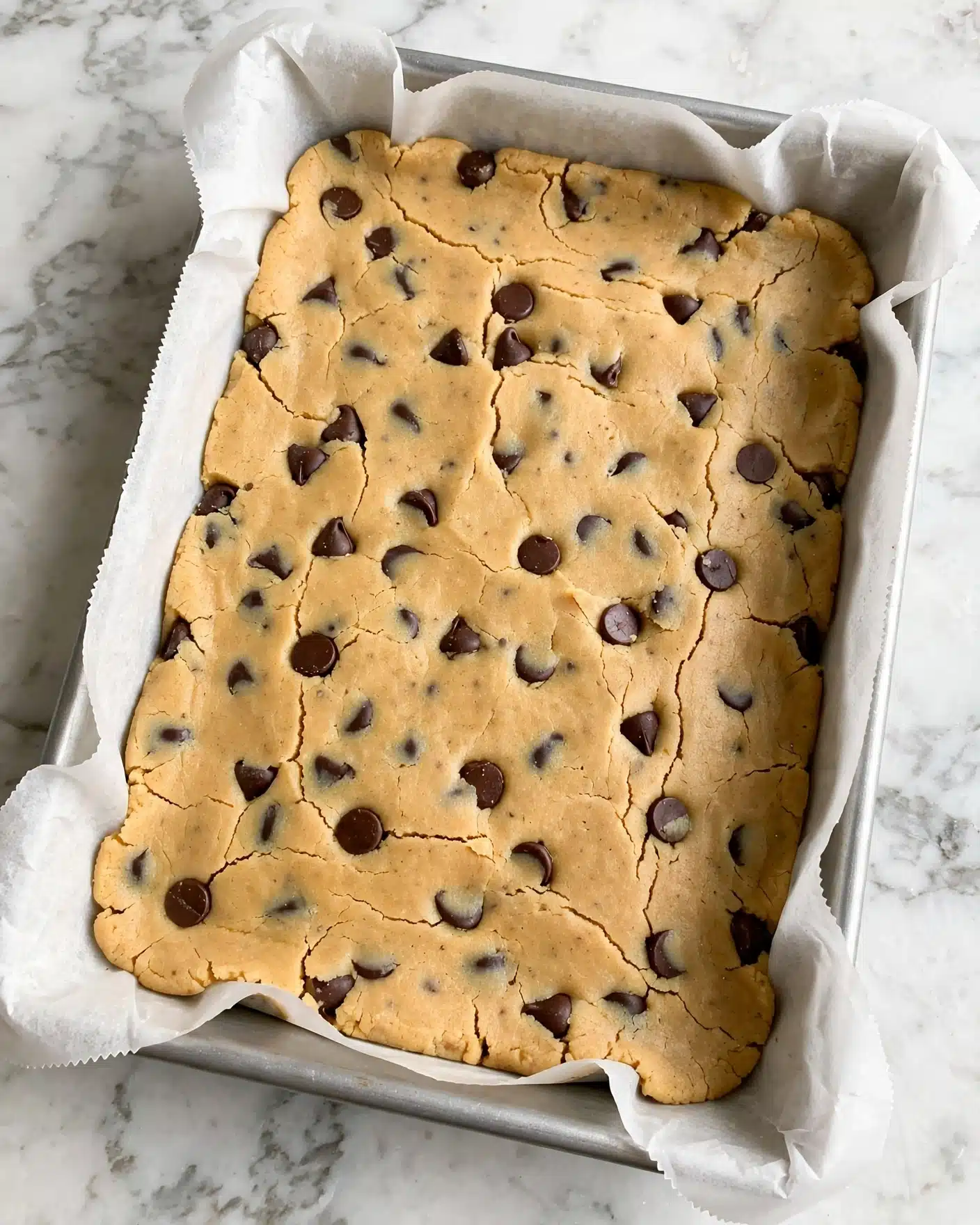 Monster Peanut Butter Cookie Bars Recipe 4 A rectangular bar of light golden cookie dough mixed with dark brown chocolate chips, pressed evenly and smoothly inside white parchment paper lining a baking pan, showing slight cracks on the surface and a firm, dense texture, all placed on a white marbled surface photo taken with an iphone --ar 4:5 --v 7
