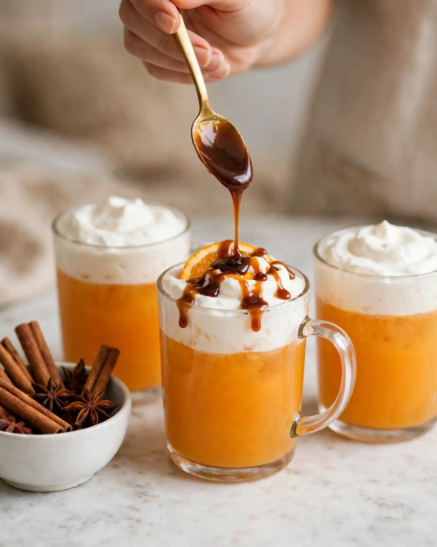 Spiced Apple Cider Recipe 5 Three clear glass mugs are filled with an orange-colored drink topped with a thick white whipped cream layer. A woman's hand holds a gold spoon above the middle mug, dripping dark caramel sauce onto the whipped cream. The whipped cream on the middle mug has a small orange slice partially visible under the caramel. The mugs sit on a white marbled surface, with a white bowl containing cinnamon sticks and star anise placed to the left. The background is soft and blurred, with warm beige tones. Photo taken with an iphone --ar 4:5 --v 7