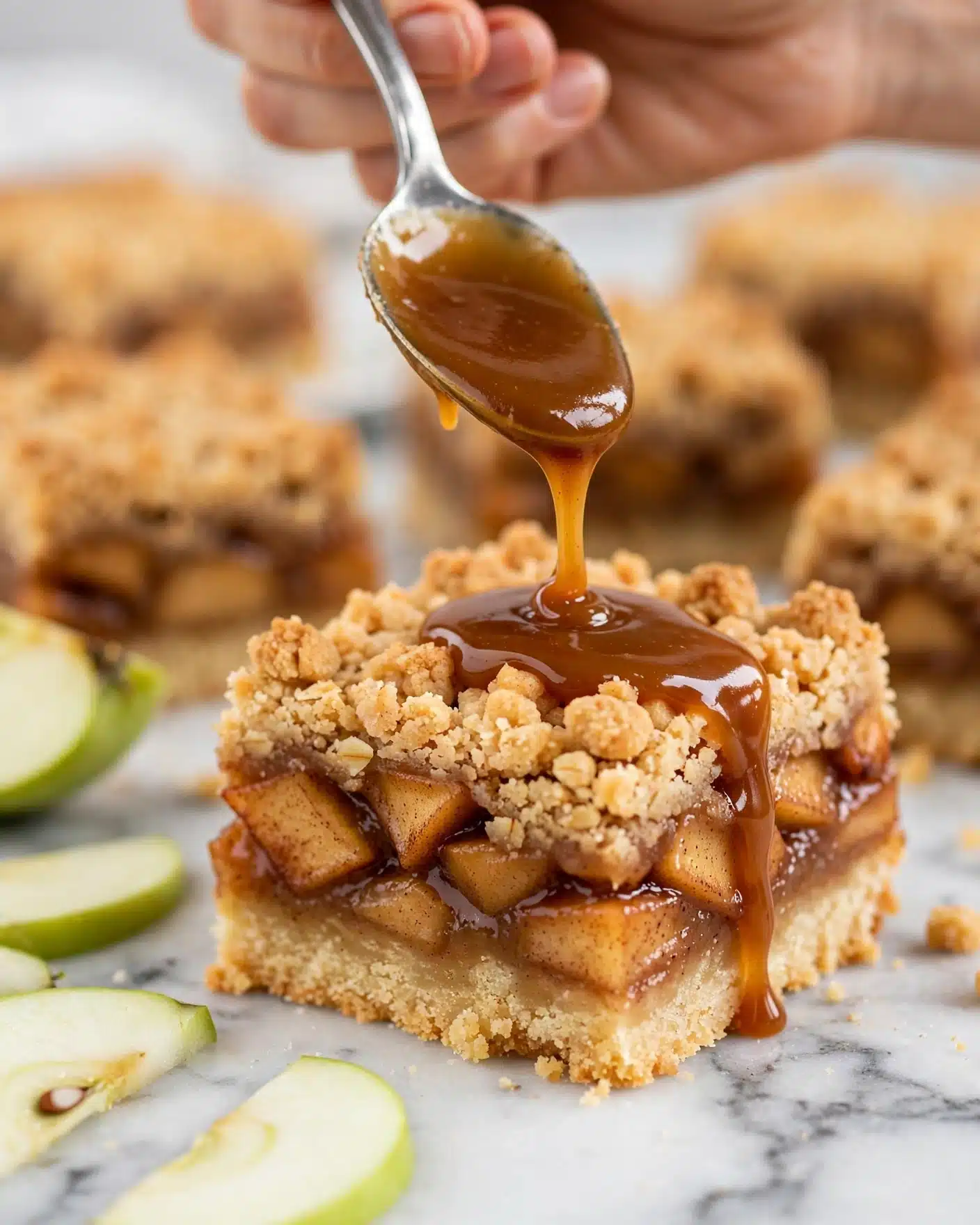 The image shows sixteen square crumb bars in a rough 4x4 layout on crumpled parchment paper atop a white marbled surface. Each bar has two visible layers: a golden brown crumbly topping textured with small uneven clumps, and a darker, sticky fruit filling layer beneath. Near the top center, there is a small glass jar filled with amber caramel sauce with a silver spoon inside. To the top right, there is a linear arrangement of five thin, pale green apple slices with a smooth texture. Two cinnamon sticks with rough bark texture lie near the top left, accompanied by a wooden-handled utensil with smooth, rounded detailing. A striped gray and white cloth is partially visible in the top right corner. photo taken with an iphone --ar 4:5 --v 7