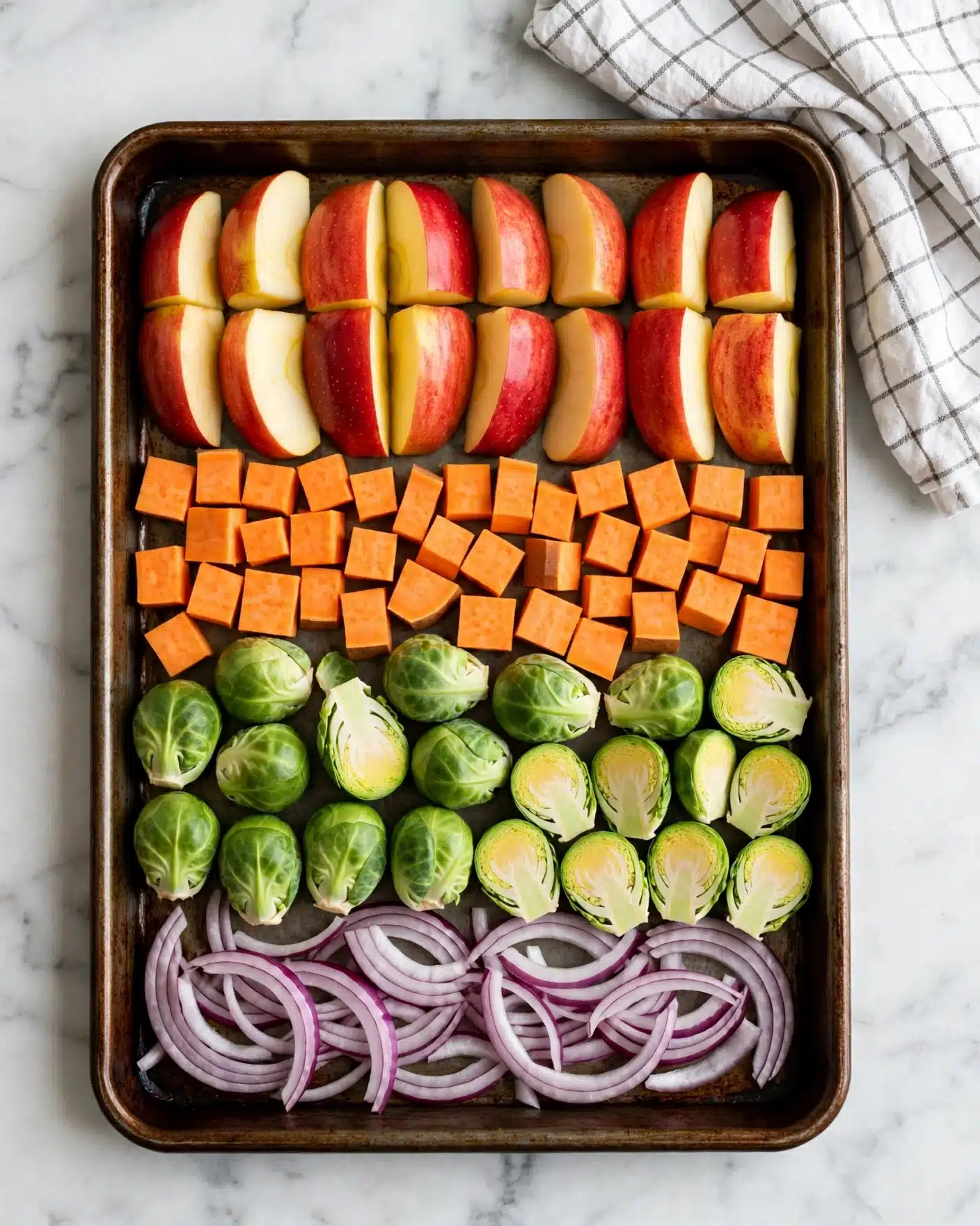The image shows a baking tray filled with four layers of colorful vegetables and fruits, arranged in neat rows from top to bottom. The top layer has red apple wedges with their skin on, showing a mix of red and pale yellow colors, the pieces are thick and arranged evenly. Below the apples, there is a layer of small orange cubes of sweet potato, cut into uniform pieces. The next row features green Brussels sprouts, some whole and some cut in half showing their tight, leafy texture inside. The bottom layer holds thin slices of purple-red onion with pale white centers, spread loosely across the tray. The tray rests on a white marbled surface, and a white checkered cloth is on the upper-right side of the image. Photo taken with an iphone --ar 4:5 --v 7