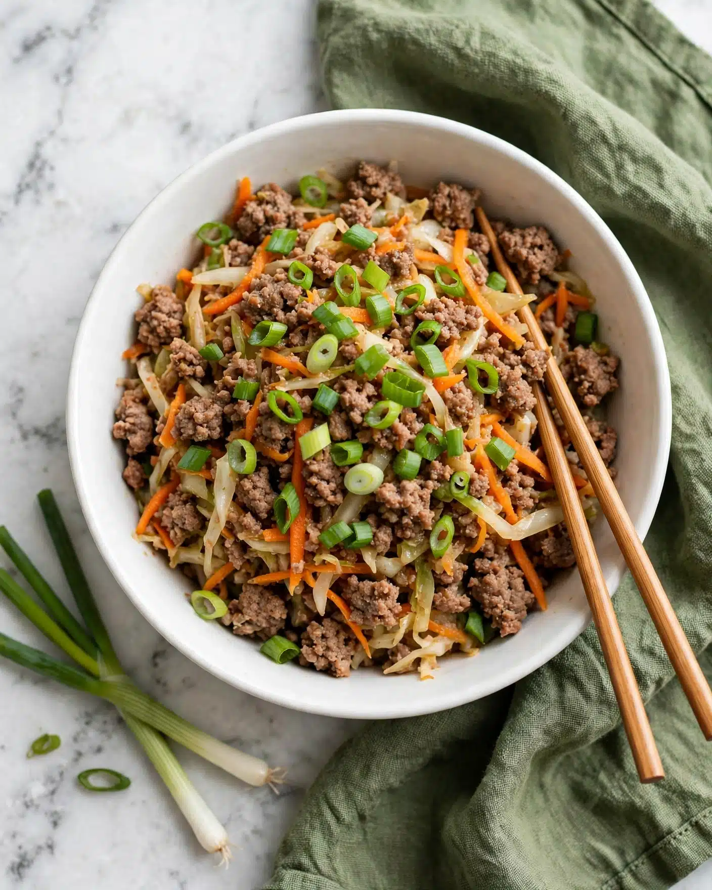 Egg Roll in a Bowl Recipe 5 A white bowl filled with a mix of ground meat and shredded vegetables, including orange carrots and light-colored cabbage, all cooked together and topped with bright green sliced scallions scattered evenly. Two wooden chopsticks rest inside the bowl on the right side. The bowl is placed on a green cloth on a white marbled surface with a few whole scallions lying nearby. photo taken with an iphone --ar 4:5 --v 7