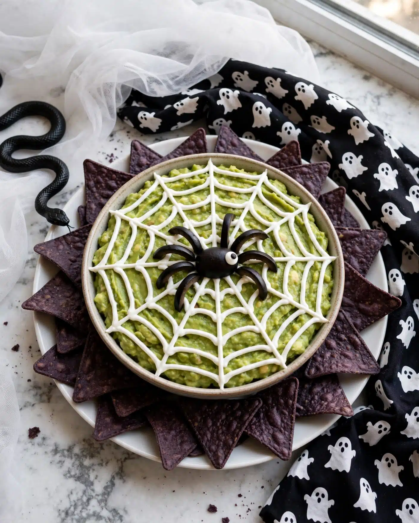 Guacamole Witch Recipe 4 A round bowl filled with green guacamole has a white sour cream spider web design on top, with the web lines spread evenly across the surface. In the center, there is a black olive spider made with sliced olives and a small white eye on one side. The bowl is placed on a white plate filled with dark purple tortilla chips around the edges. The background has a white marbled texture covered partly with black and white cloth featuring ghost patterns and some white sheer fabric. A black plastic snake is also positioned on the top left side near the plate. photo taken with an iphone --ar 4:5 --v 7