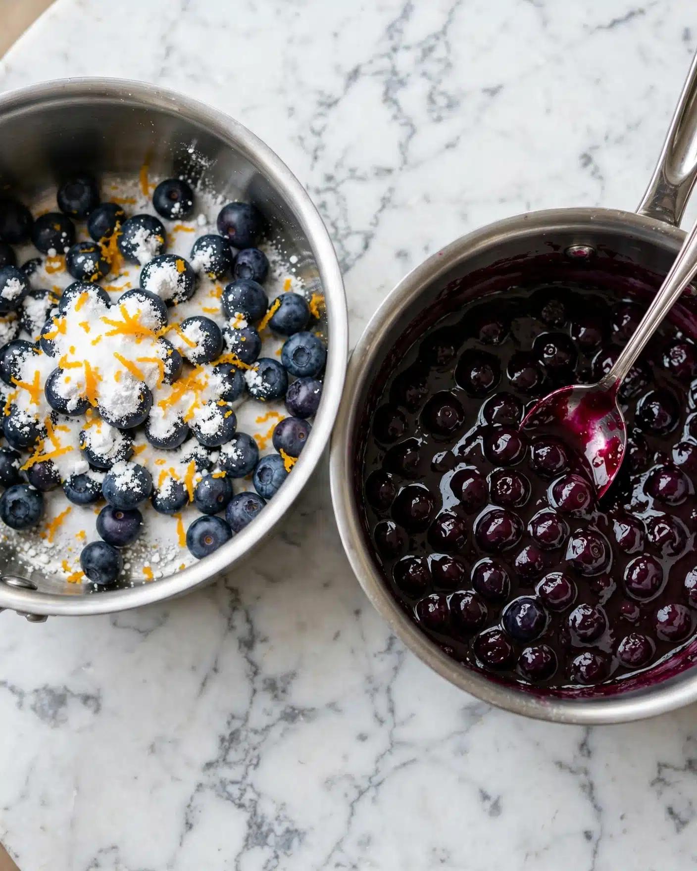 Gluten-Free Blueberry Pop Tarts Recipe 4 The image shows two metal saucepans side by side on a white marbled surface. The left saucepan contains fresh dark blueberries piled in the pot with white sugar, white powdered sugar, and bright orange zest scattered on top, all unmixed. The right saucepan shows the cooked blueberry mixture transformed into a thick, dark purple sauce with whole blueberries still visible, a shiny texture, and a silver spoon resting inside. photo taken with an iphone --ar 4:5 --v 7