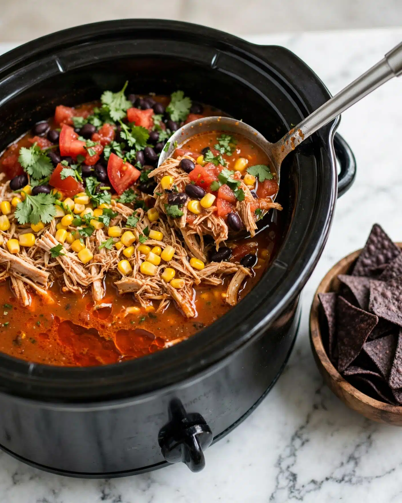 Slow Cooker Chicken Tortilla Soup Recipe 5 A black slow cooker filled with chunky soup showing three main layers: a reddish-orange broth base with a slightly oily texture, a middle layer of shredded light brown chicken and bright yellow corn kernels, and a top layer of fresh green cilantro leaves scattered along with chunks of red tomatoes and black beans throughout. A silver ladle dipping into the soup reveals the soup’s chunky consistency, all placed on a white marbled surface with a wooden bowl of dark blue tortilla chips nearby. Photo taken with an iphone --ar 4:5 --v 7