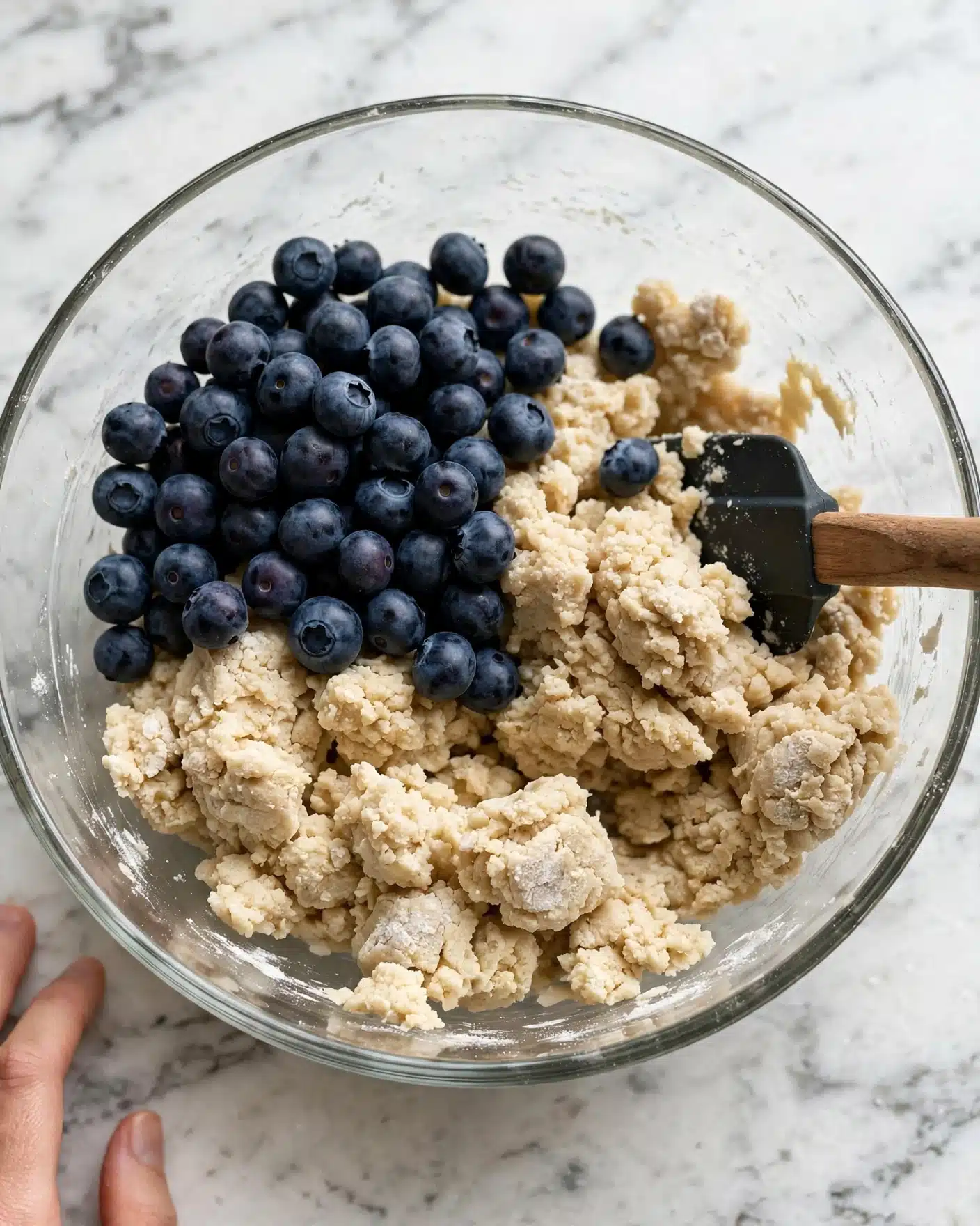 Low Fat Blueberry Scones Recipe 4 A clear glass bowl placed on a white marbled surface contains a mix of light beige dough and a pile of fresh dark blue blueberries. The dough is chunky and uneven, covering roughly half of the bowl's bottom and sides. A black spatula with a wooden handle is partially buried in the dough on the right side of the bowl. A woman's hand is just touching the bottom left corner of the image, resting on the white marbled surface nearby. photo taken with an iphone --ar 4:5 --v 7
