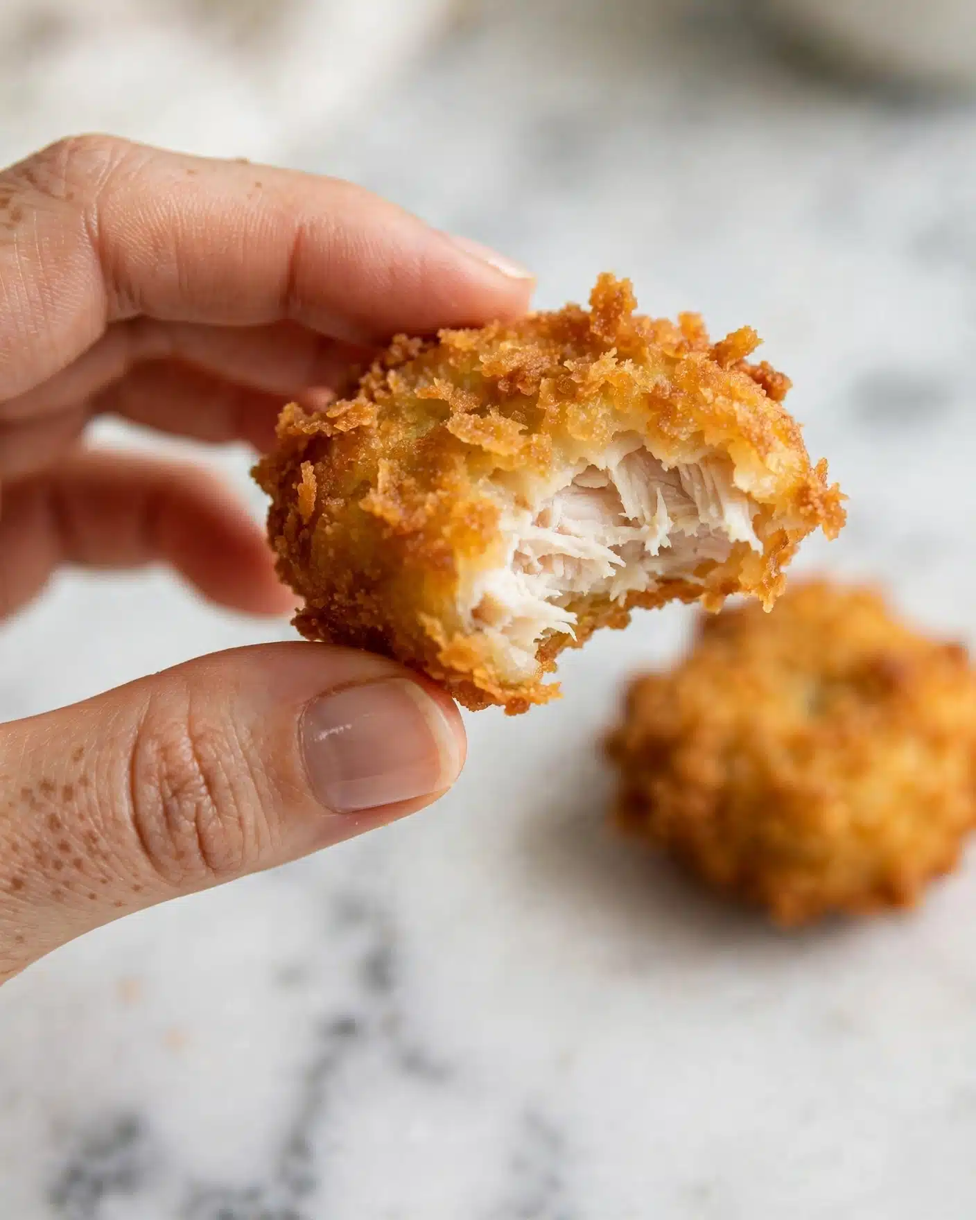 Baked Chicken Nuggets Recipe 5 A close-up image shows a woman's hand holding a half-eaten golden-brown crispy battered nugget. The nugget has an outer crunchy layer with a rough texture, while the inside is soft and white. The background features a blurred nugget on a white marbled surface. Photo taken with an iphone --ar 4:5 --v 7
