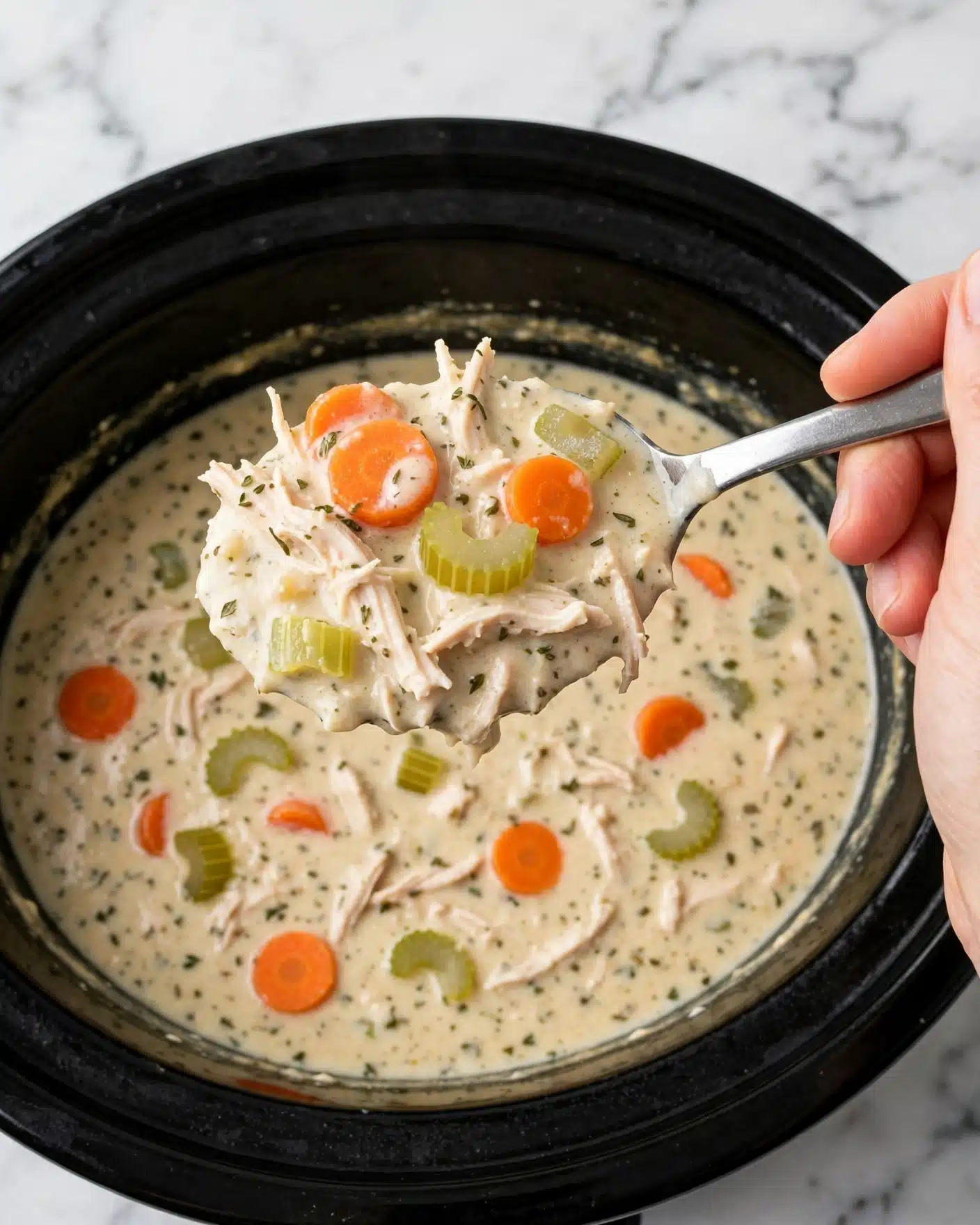 Creamy Crockpot Chicken Wild Rice Soup Recipe 5 A close-up view of a thick, creamy soup served in a large black slow cooker. The soup has three main layers: a light beige creamy base with a smooth texture, with small bits of herbs scattered throughout; floating irregular pieces of shredded white chicken; and bright orange carrot slices and pale green celery pieces spread evenly in the soup. The spoon, held by a woman's hand, lifts a serving showing all these layers and colors clearly. The background is a white marbled texture. photo taken with an iphone --ar 4:5 --v 7