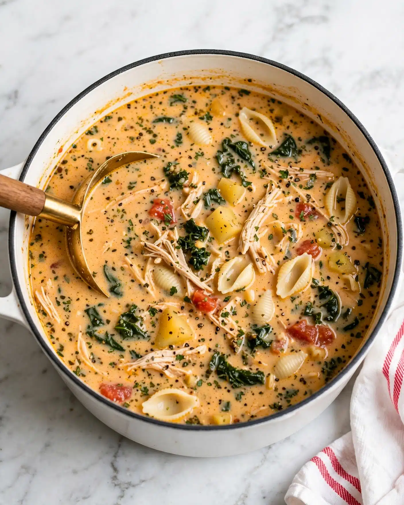 Creamy Marry Me Chicken Soup (30-Minute Recipe!) Recipe 4 The image shows a creamy soup inside a round white pot with a dark rim. The soup has multiple layers visible: at the base, there is a thick light orange broth with specks of herbs and spices. Mixed within are shredded light beige chicken pieces and small white pasta shells. There are also dark green leafy vegetables scattered throughout, adding texture and color contrast. Small chunks of yellow potato and some red tomato bits float in the soup. A golden ladle with a wooden handle is partially dipped into the soup on the left side. The pot sits on a white marbled surface with a part of a white cloth with red stripes visible on the lower right side. photo taken with an iphone --ar 4:5 --v 7