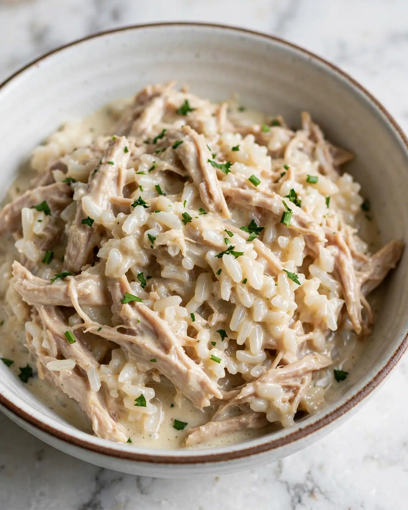 Creamy Chicken and Rice Casserole Recipe 5 A close-up view of a creamy dish served in a white bowl with a thin brown rim, placed on a white marbled surface. The dish has a mix of soft, light beige shredded meat and slightly sticky rice, both coated in a light creamy sauce. The rice grains are small and well-cooked, tightly packed but still visible within the sauce. Small bits of fresh green herbs are sprinkled evenly over the top, adding a touch of color contrast to the creamy beige mix. Photo taken with an iphone --ar 4:5 --v 7