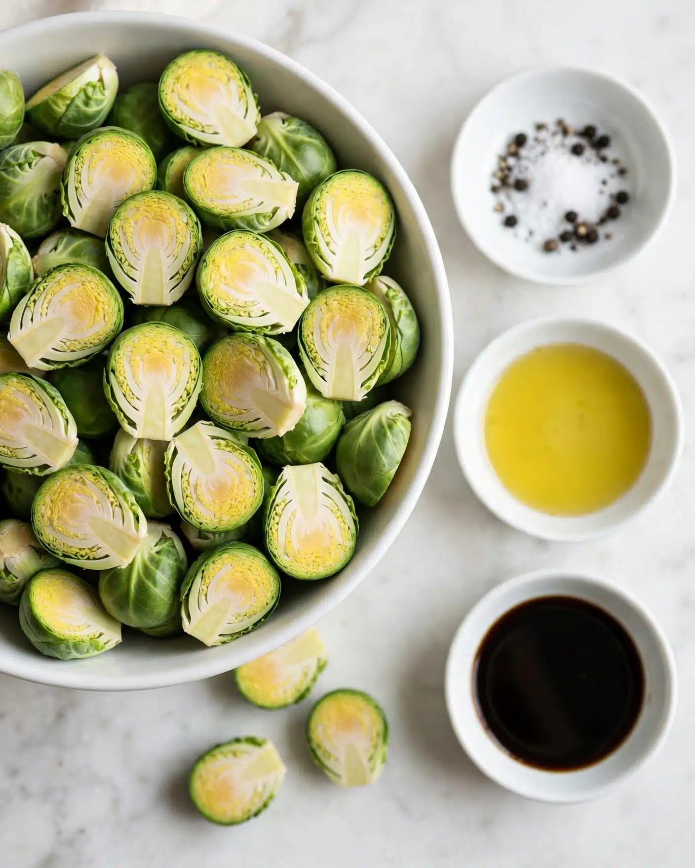 Balsamic Roasted Brussels Sprouts Recipe 4 A large white bowl filled with halved Brussels sprouts showing their light green inner layer and darker green outer leaves, sitting on a white marbled surface. To the right, there are three smaller white bowls; the top one holds salt and black pepper, the middle one contains a pale yellow liquid, likely oil, and the bottom one has a dark liquid, possibly soy sauce. The colors contrast well with the green Brussels sprouts and light background, creating a fresh and simple composition. photo taken with an iphone --ar 4:5 --v 7