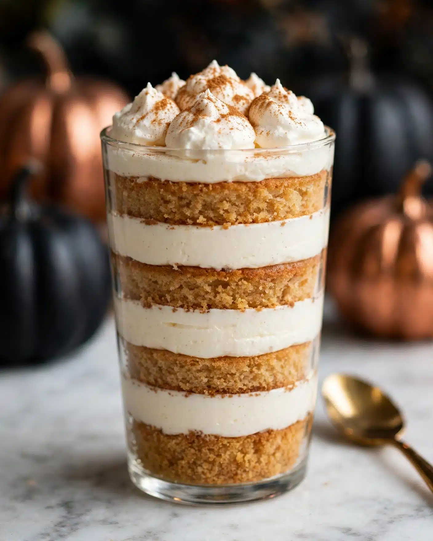 The image shows a clear glass filled with five visible layers, starting with a light brown cake layer at the bottom, followed by a thick white cream layer, then another light brown cake layer, a second thick white cream layer, and topped with dollops of white whipped cream sprinkled with light brown spice powder. The glass sits on a gray cloth, with a gold spoon featuring a white handle resting in front on a white marbled surface. The background is softly blurred with dark, neutral tones. Photo taken with an iphone --ar 4:5 --v 7