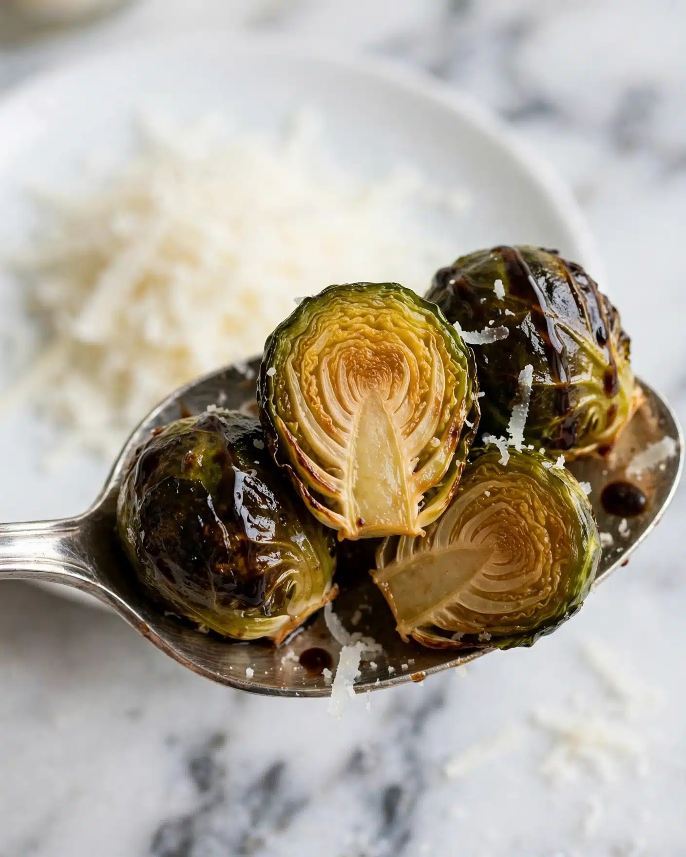 Balsamic Roasted Brussels Sprouts Recipe 5 A close-up of a silver spoon holding several roasted Brussels sprouts, some whole and some cut in half showing their soft, golden-brown interiors. The sprouts are shiny with dark char marks and a drizzle of dark sauce on top. There are thin white shavings, likely cheese, scattered over the sprouts on the spoon and piled in the background on a white plate. The background has a white marbled texture. photo taken with an iphone --ar 4:5 --v 7