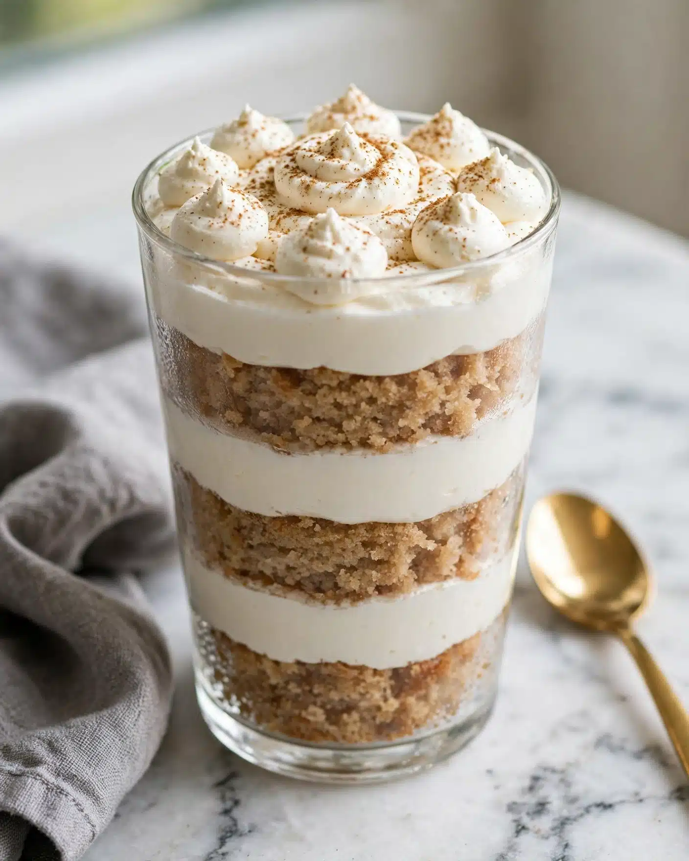 The image shows a clear glass filled with multiple layers of dessert stacked neatly. The bottom layer is a golden-brown cake, followed by a thick white cream layer, then another cake layer, and another cream layer. This pattern repeats twice more, making a total of five cake layers and four cream layers. On top, there are several dollops of whipped cream sprinkled with a light dusting of cinnamon. The glass is placed on a white marbled surface, with a gold spoon nearby and blurred black and copper decorative pumpkins in the background. Photo taken with an iphone --ar 4:5 --v 7