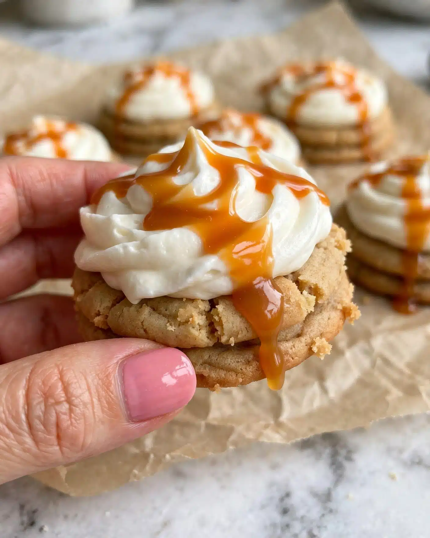 A close-up of a cookie held by a woman's hand with pink nail polish, showing three visible layers: a light brown soft cookie base, a thick swirl of white creamy frosting on top, and a shiny drizzle of orange caramel sauce flowing over the frosting and slightly dripping down the side. The background shows more cookies with the same layers on a crinkled beige parchment paper over a white marbled surface. photo taken with an iphone --ar 4:5 --v 7