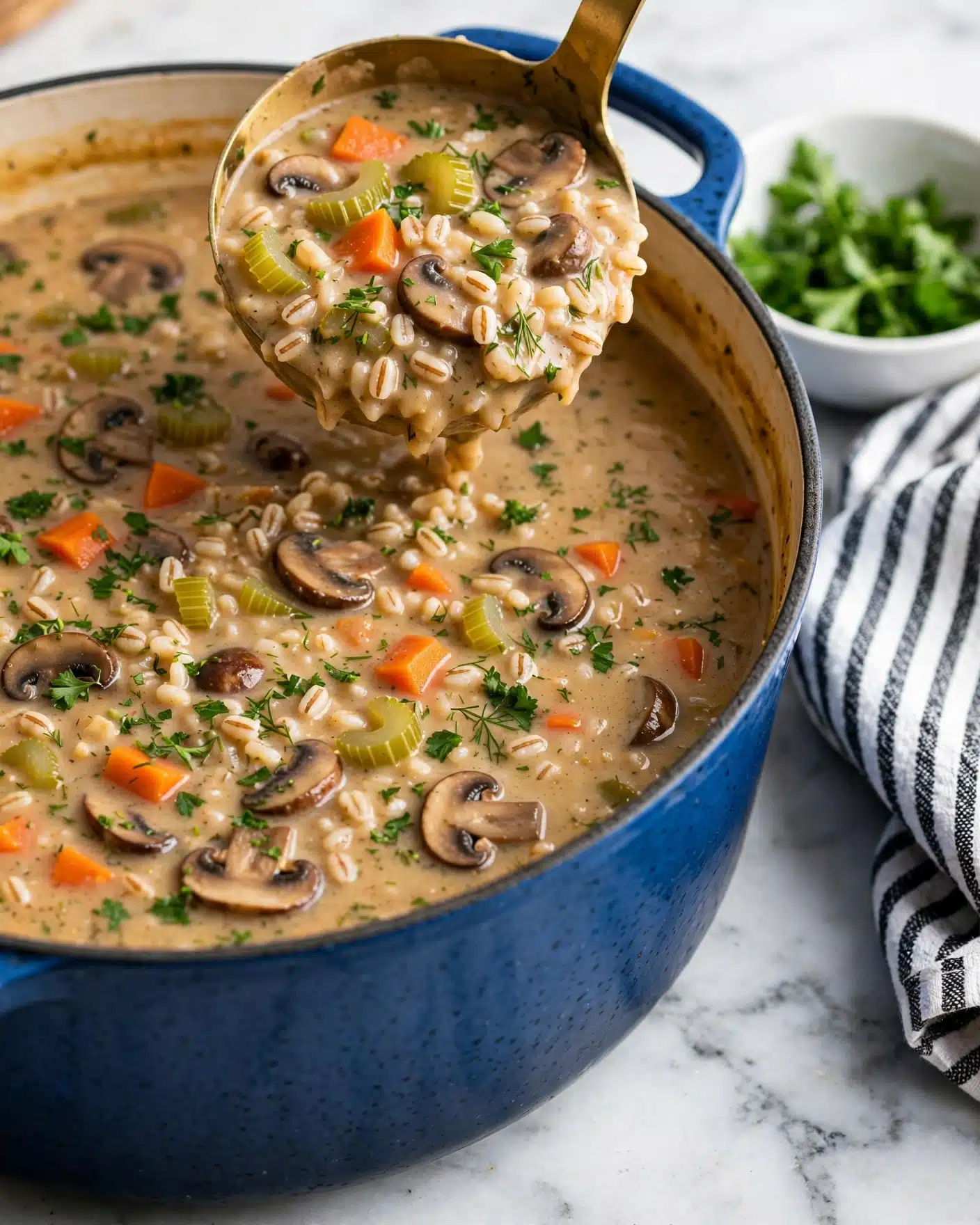 Mushroom Barley Soup Recipe 5 The image shows a close-up of a large blue pot filled with thick, creamy soup. The soup has visible layers of chopped orange carrots, green celery, sliced brown mushrooms, and small grains that seem soft and plump. The soup is a warm light brown color with mixed-in fresh green herbs sprinkled evenly on top. A golden ladle is partially lifted inside the pot, filled with a generous scoop of the same soup, showing the chunky vegetables and grains clearly. The pot sits on a white marbled surface next to a small white bowl of fresh green herbs and a white and black striped cloth nearby. The photo taken with an iphone --ar 4:5 --v 7