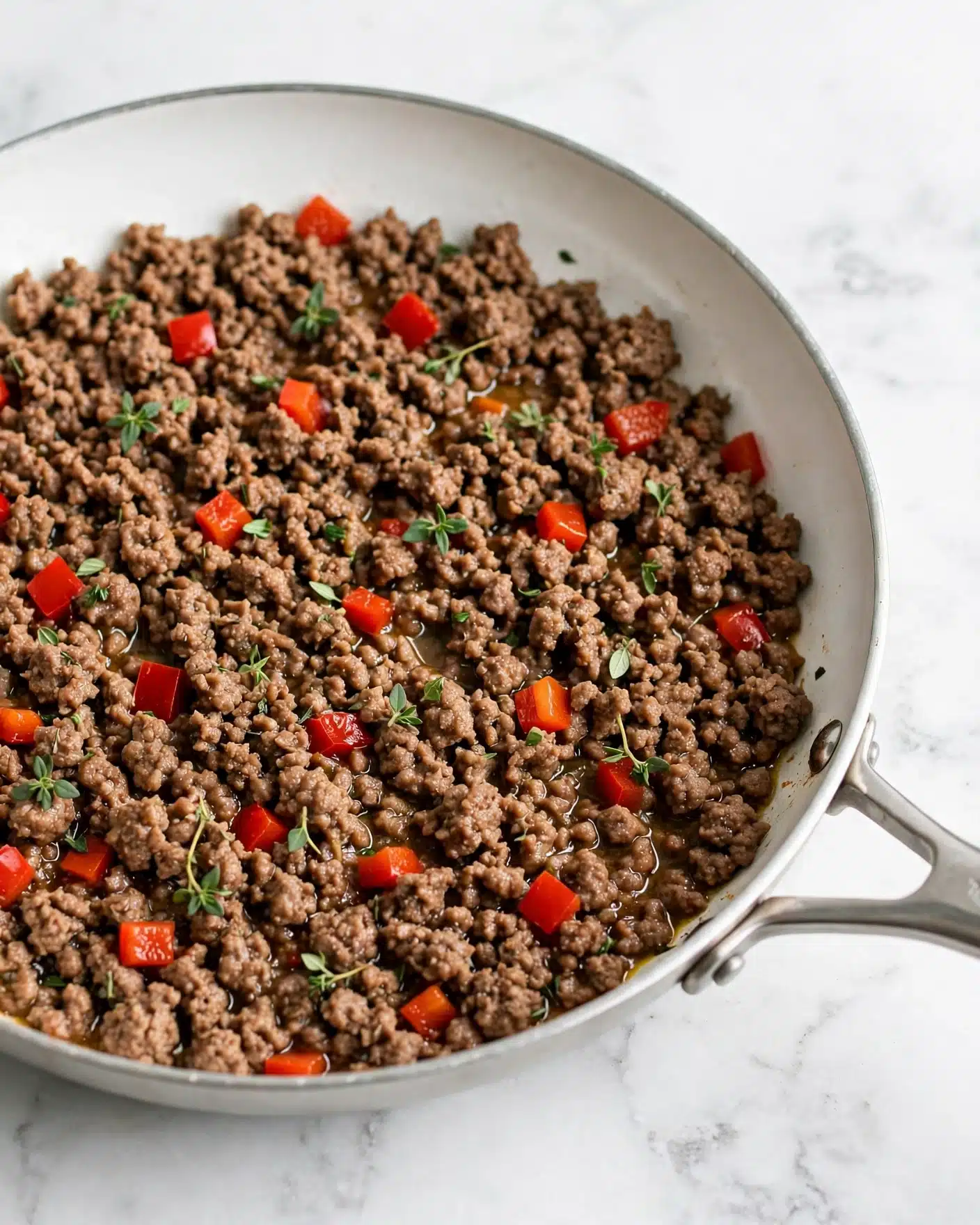 This image shows a white pan filled with cooked ground meat that is brown with a slightly oily texture. Small pieces of bright red bell pepper are scattered throughout the meat, adding color contrast. Tiny green herb leaves are sprinkled on top, giving a fresh touch. The white marbled surface under the pan is clean and smooth, highlighting the dish. Photo taken with an iphone --ar 4:5 --v 7