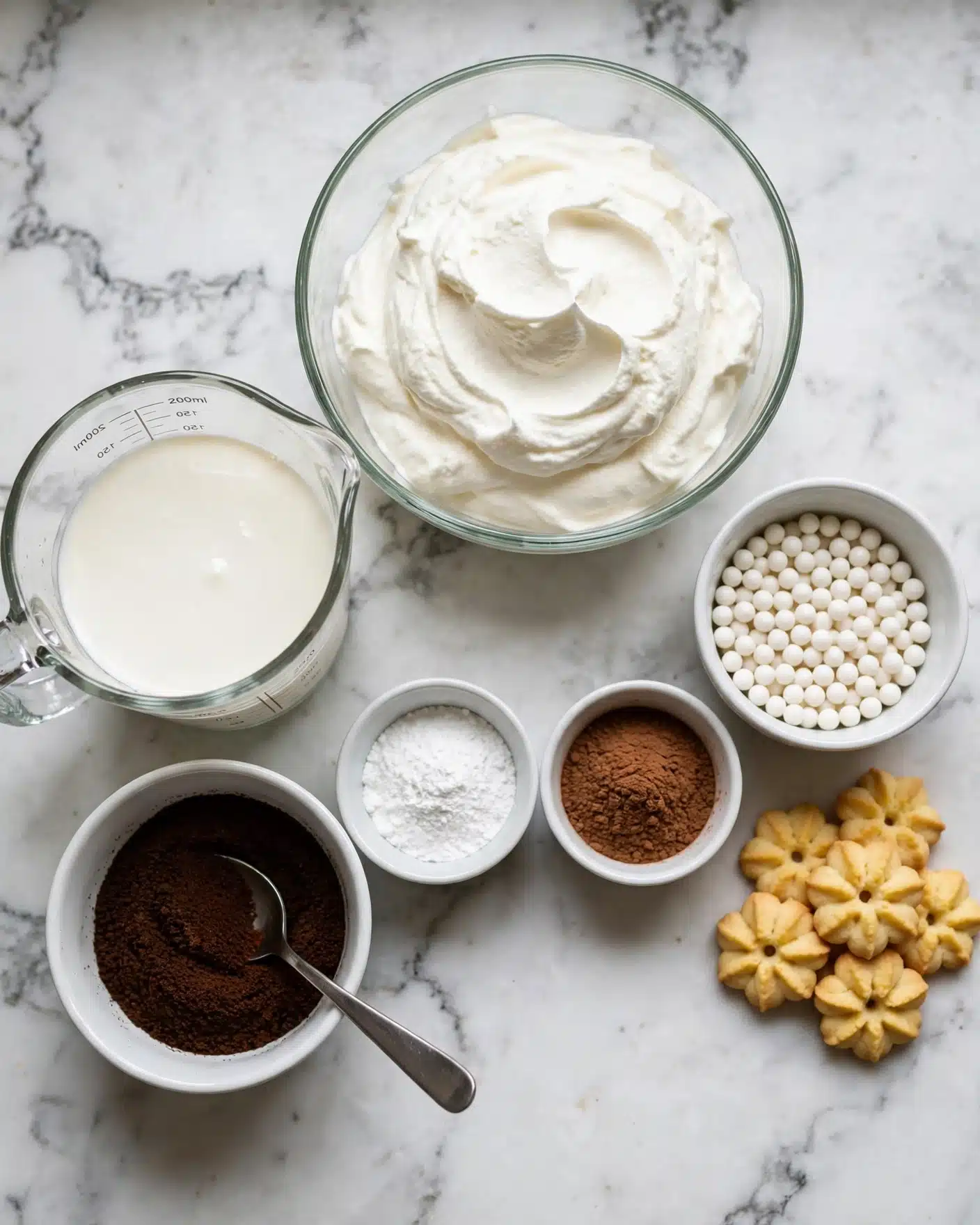 Copycat Disney Grey Stuff Recipe 4 The image shows several ingredients for a dessert arranged on a white marbled surface. There is a clear glass bowl filled with thick white whipped cream in the center top. To the left is a transparent measuring cup with cream. Below that is a round white bowl with dark brown coffee grounds and a silver spoon inside. Next to it are two small round bowls; one with white powder and the other with a brown powder. On the right side, there is a small white bowl filled with white candy pearls, and nearby are yellowish flower-shaped cookies with holes in the middle arranged in a small cluster. Photo taken with an iphone --ar 4:5 --v 7