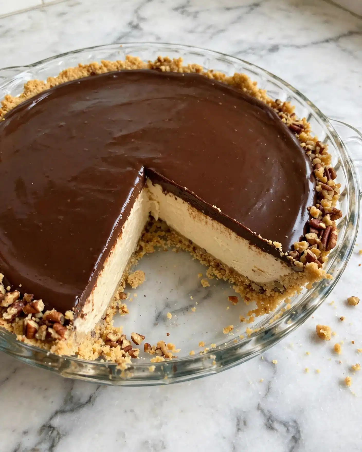 Peanut Butter Pretzel Pie Recipe 5 A clear glass pie dish holds a three-layer dessert placed on a white marbled surface. The bottom layer is a crumbly, light golden crust with visible crushed nuts pressed firmly along the edge and base. Above the crust is a thick, creamy beige middle layer, smooth in texture and solid in shape. The top layer is a shiny, dark brown chocolate spread spread evenly, covering the entire surface except the crust edge. A missing wedge reveals all three layers clearly. Photo taken with an iphone --ar 4:5 --v 7