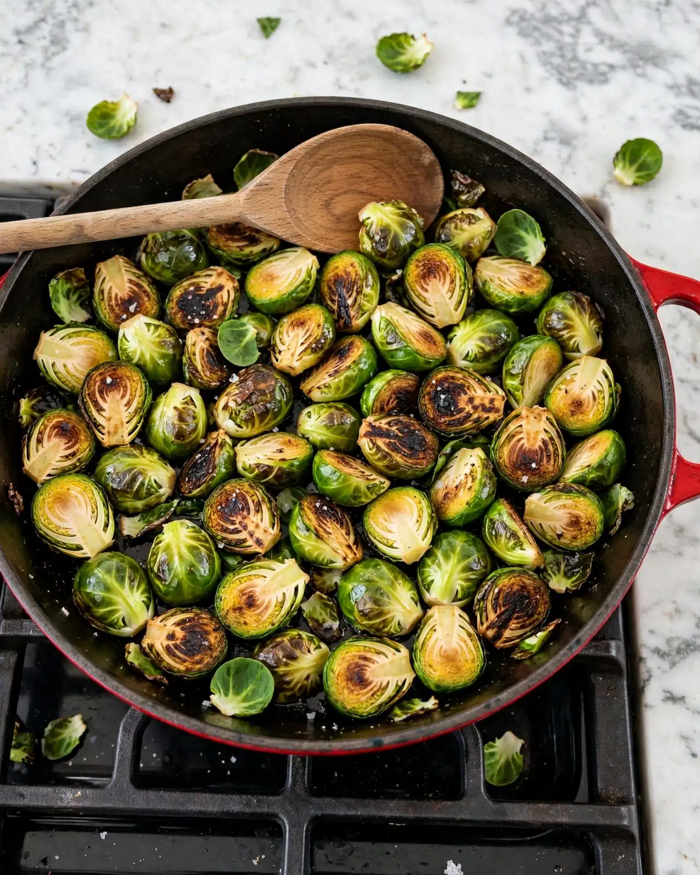 The image shows a red cast iron pan on a black stove grate, filled with bright green Brussels sprouts that are halved and slightly browned on the cut sides, showing a mix of smooth and charred textures. Some loose Brussels sprout leaves are scattered among the pieces. A wooden spoon is resting inside the pan, positioned over the sprouts with a visible round and smooth surface. The surrounding surface is a white marbled texture. Photo taken with an iphone --ar 4:5 --v 7