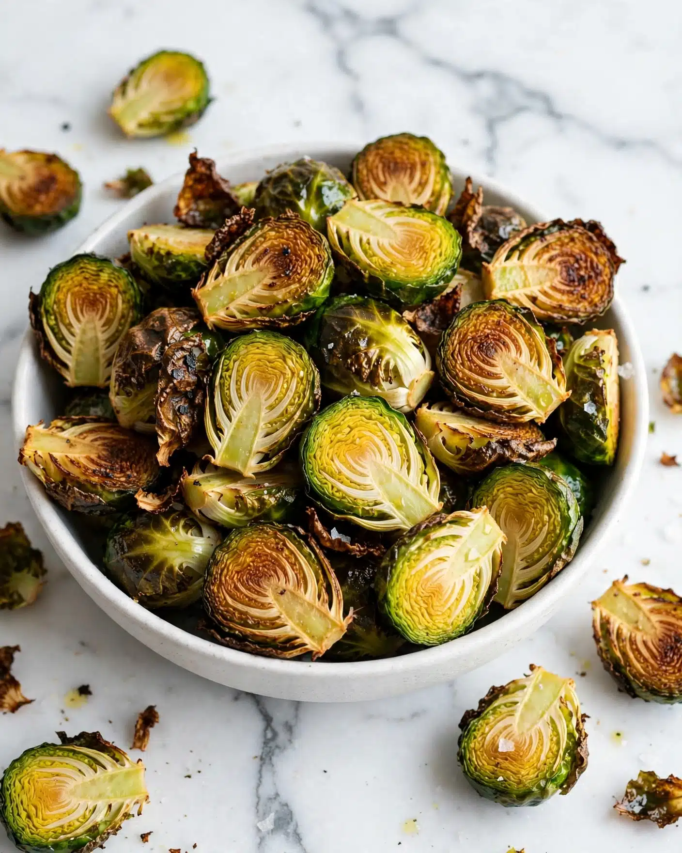 A white bowl filled with golden-brown roasted Brussels sprouts, each piece cut in half showing a caramelized, slightly crispy surface with bright green and yellow-green inner layers. The roasted edges are dark brown and textured, contrasting with the softer, lighter green parts inside. The bowl sits on a wooden surface replaced by a white marbled background. photo taken with an iphone --ar 4:5 --v 7