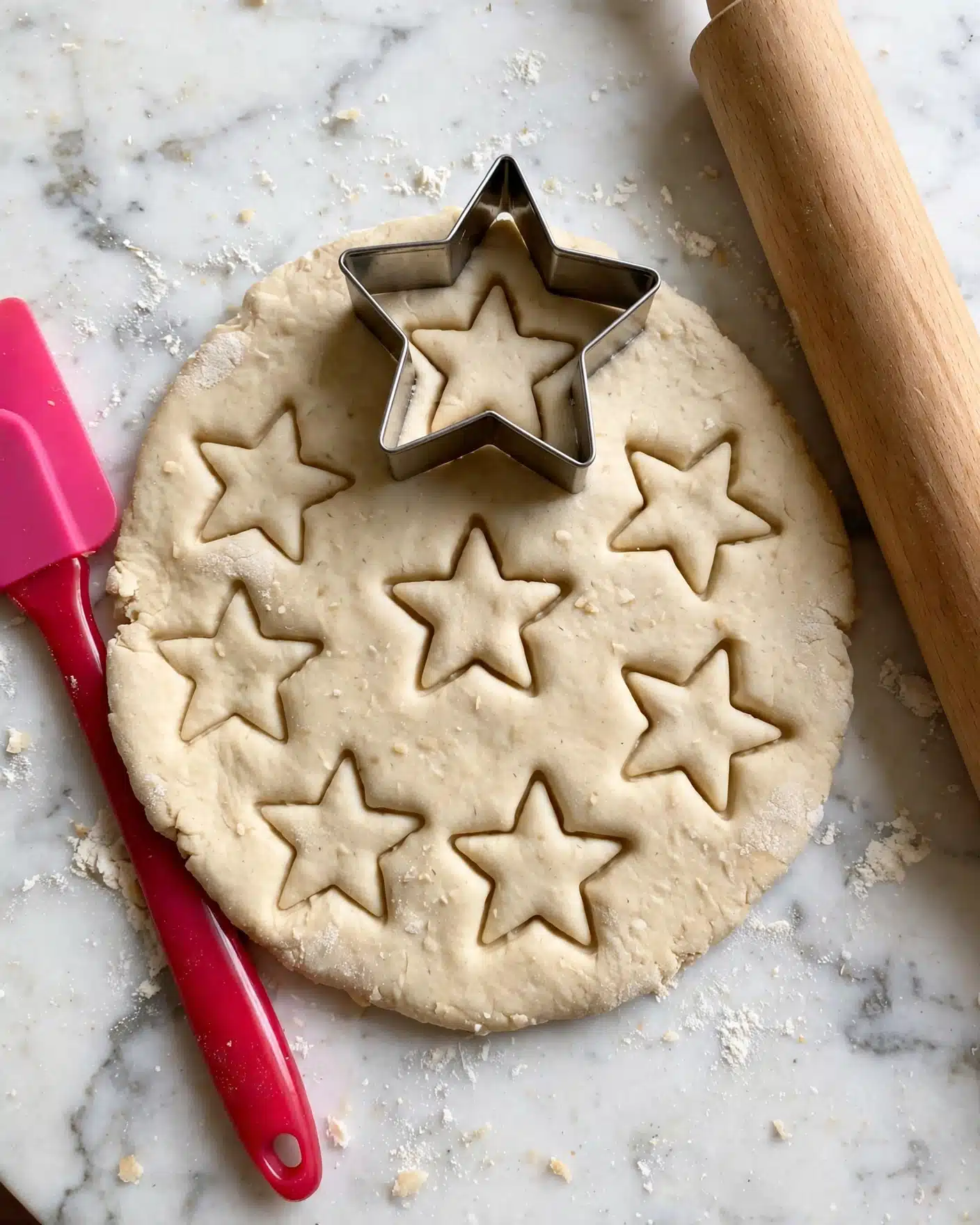 Holiday Sugar Cookies Recipe 4 A flat piece of light beige dough laid out on a white marbled surface, with seven star shapes pressed gently into the dough, leaving clear star outlines and depth. To the left of the dough is a bright pink silicone spatula with a red handle, resting partially on the surface. Above the dough is a silver star-shaped metal cookie cutter, positioned with one star already shaped in the dough beneath it. To the right side of the image is a light brown wooden rolling pin angled diagonally. photo taken with an iphone --ar 4:5 --v 7