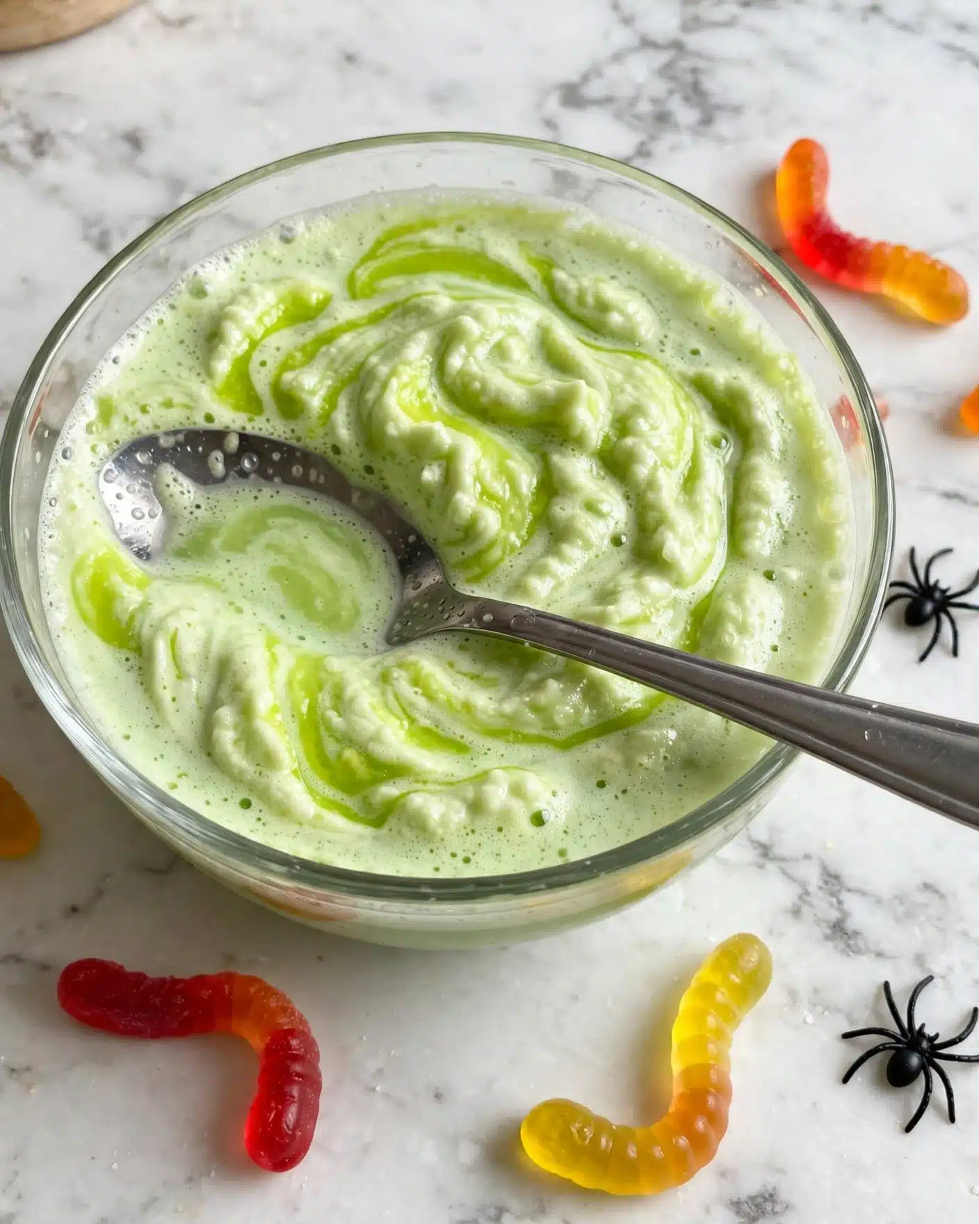 Easy Spooky Non-Alcoholic Punch for Halloween Recipe 5 A clear glass bowl filled with a light green, foamy dessert with a creamy texture, presented on a white marbled surface covered with an orange cloth with black stitch lines. Inside the bowl, there are two visible gummy worms, colored red, yellow, and orange, placed on top of the foamy layer. There is a small black plastic spider sitting near the edge of the bowl, close to a silver spoon partially visible in the foreground. The dessert has a bubbly, airy appearance with a few darker green patches on the surface. Photo taken with an iphone --ar 4:5 --v 7