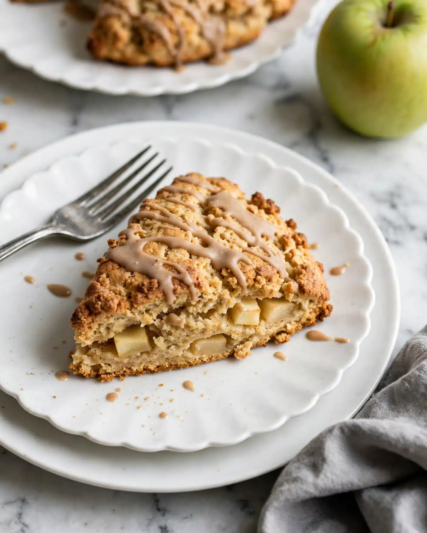 Apple Cinnamon Diced Apple Scones with Vanilla Glaze Recipe 5 A single slice of pie sits on a white plate with scalloped edges, layered on top of another plain white plate. The pie slice has three visible layers: a crumbly golden brown crust at the bottom, a lighter beige filling in the middle, and a top layer covered with a smooth, light brown glaze with drizzle patterns. Small splashes of the glaze are on the plate around the pie. A silver fork rests on the left side of the plate. Behind this plate, a partial view of another slice of pie on a similar white scalloped plate is visible, along with a whole green apple on a white marbled surface. A folded grey cloth is in the bottom right corner. photo taken with an iphone --ar 4:5 --v 7
