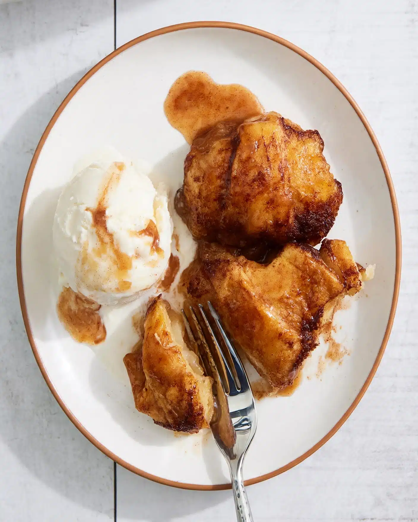 Easy Crescent Apple Dumplings Recipe 5 A white plate with a thin brown rim holds three pieces of golden-brown cooked dessert with a caramel-like sauce on top. To the left side of the plate is a round scoop of white ice cream with some of the sauce drizzled over it. The dessert pieces appear soft and slightly crispy on the outside, with one piece partially broken apart showing a tender inside. A silver fork is placed near the bottom center with its prongs holding the smaller broken piece. The plate is resting on a white marbled surface. photo taken with an iphone --ar 4:5 --v 7