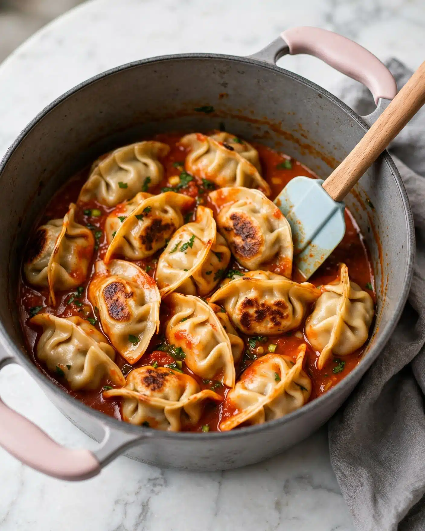 A large gray pot with light pink handles holds many dumplings in a red-orange sauce with small bits of green herbs visible. The dumplings are folded and have a pale golden color with some parts darker where they cooked more. A light blue spatula with a wooden handle is inside the pot resting on the dumplings. The pot sits on a white marbled surface with a gray cloth partially under the right side of the pot. photo taken with an iphone --ar 4:5 --v 7