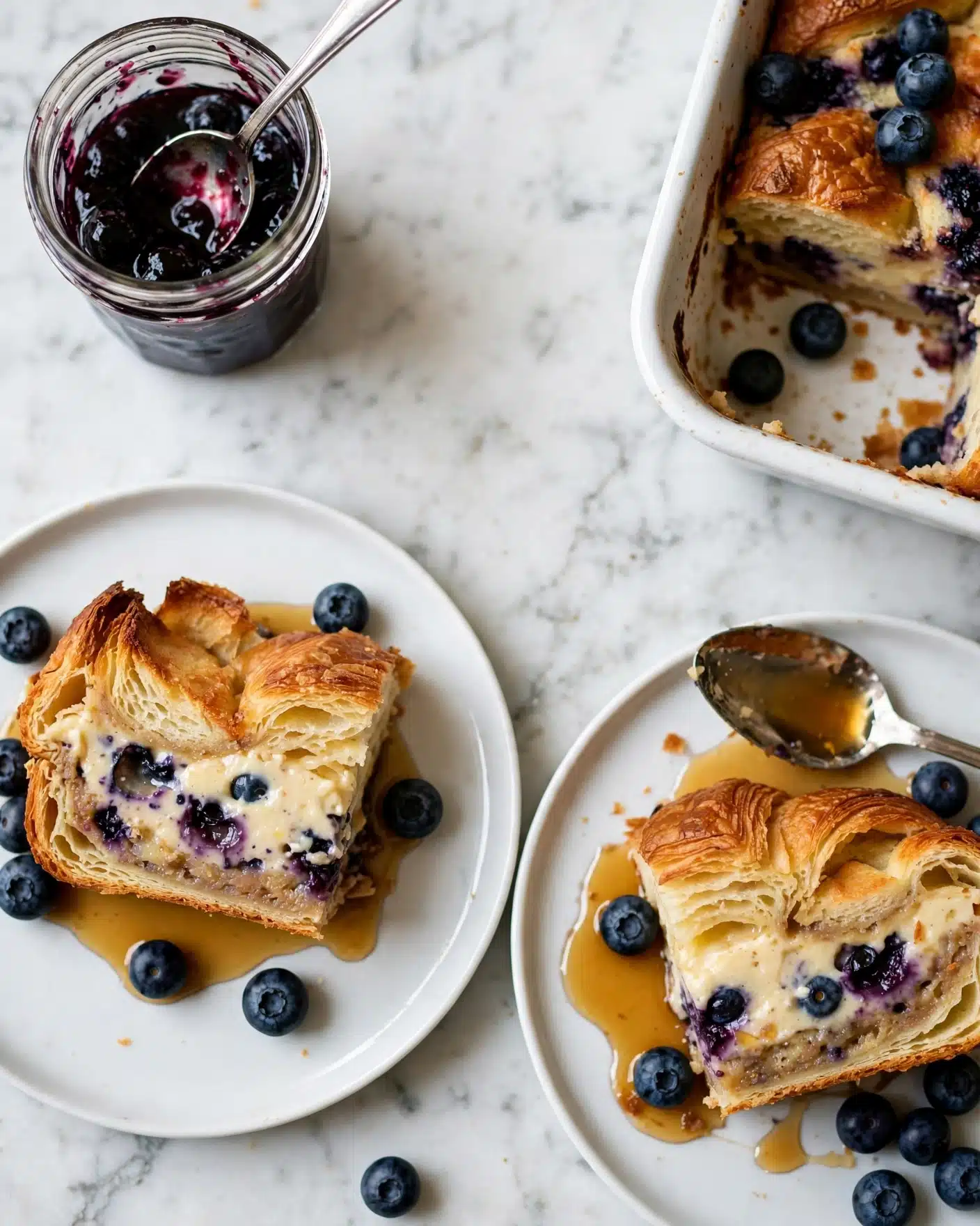 Baked Berry and Cream Cheese Croissant French Toast Recipe 5 The image shows two white plates, each with a piece of baked croissant bread pudding. Each slice has three visible layers: a top golden brown, crispy and flaky croissant layer, a creamy middle layer with a light beige color mixed with darker blueberry spots, and a bottom layer that looks soft and slightly browned. Blueberries are scattered on the plates and mixed inside the pudding. A drizzle of amber syrup pools around the food. A silver spoon rests on one plate with syrup on its bowl. Part of a white baking dish with more croissant bread pudding topped with blueberries is on the right, and a jar of dark blueberry jam with a silver spoon inside is on the top left. The background is a white marbled texture. photo taken with an iphone --ar 4:5 --v 7