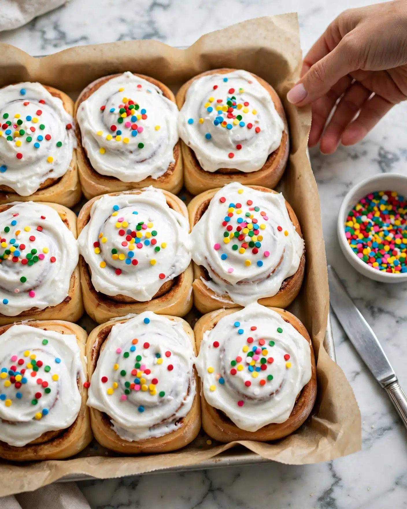 Cereal Milk Cinnamon Rolls Recipe 5 Six cinnamon rolls are arranged on a piece of parchment paper on a white marbled surface. Each roll has two layers: a golden-brown bread base with a soft, fluffy inside showing colorful bits mixed in, and a thick layer of white icing on top, sprinkled with bright, multicolored crunchy cereal pieces. One roll in the front has a bite taken out, revealing its soft texture and colorful bits inside. To the right side on the surface, there is a silver knife with some white icing on it. The background is softly blurred, emphasizing the cinnamon rolls. Photo taken with an iphone --ar 4:5 --v 7
